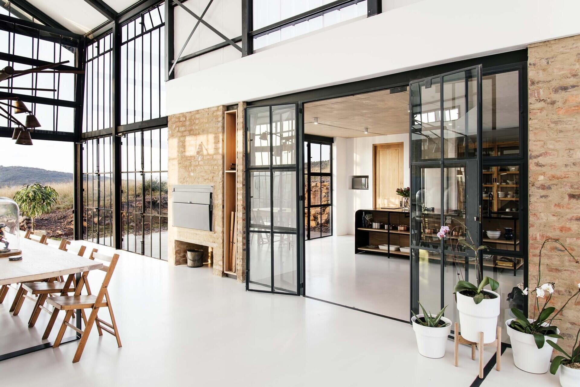 Black-framed glass doors connect the conservatory to the kitchen, where a black island becomes the focal point. Open wood shelving beneath it displays everyday items like pots, pans, and serving dishes, turning storage into part of the design.