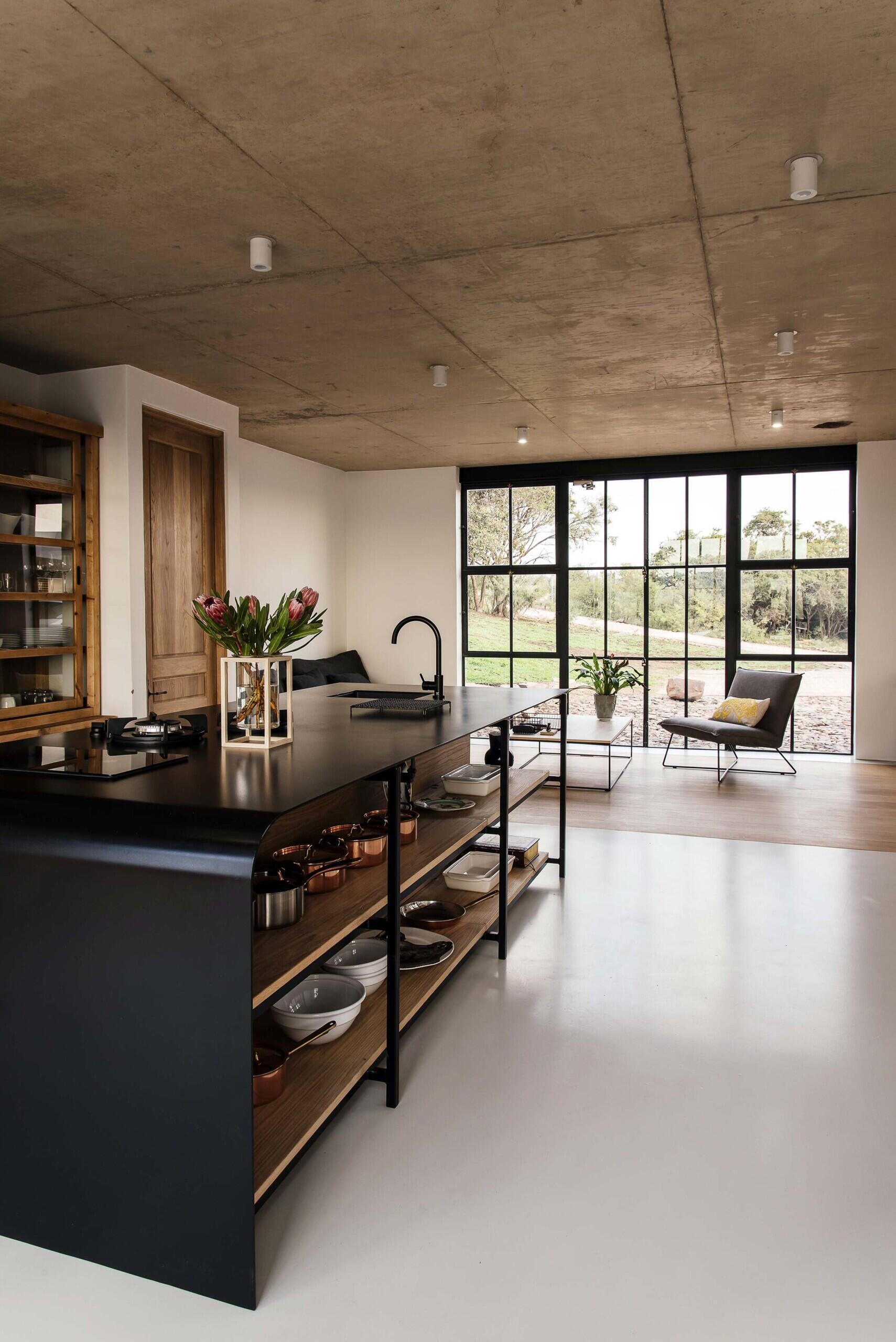 Black-framed glass doors connect the conservatory to the kitchen, where a black island becomes the focal point. Open wood shelving beneath it displays everyday items like pots, pans, and serving dishes, turning storage into part of the design.