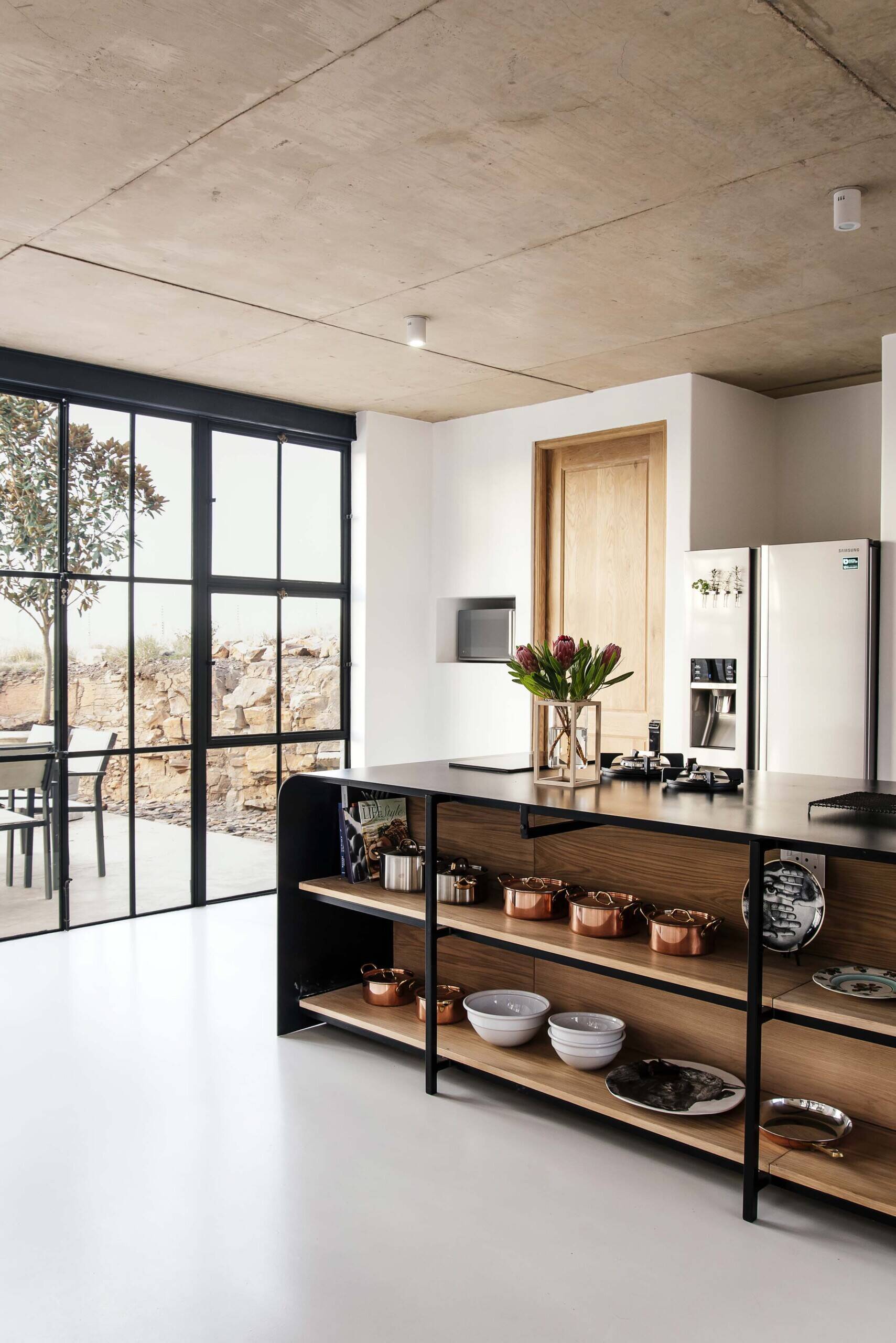 Black-framed glass doors connect the conservatory to the kitchen, where a black island becomes the focal point. Open wood shelving beneath it displays everyday items like pots, pans, and serving dishes, turning storage into part of the design.