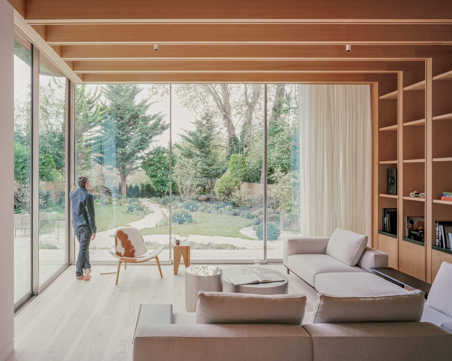 A sunlit living space steps down into the garden, using level changes and natural light to define the layout. The wood shelving in the living room adds a quieter layer to the space, balancing out the openness created by the glass walls.