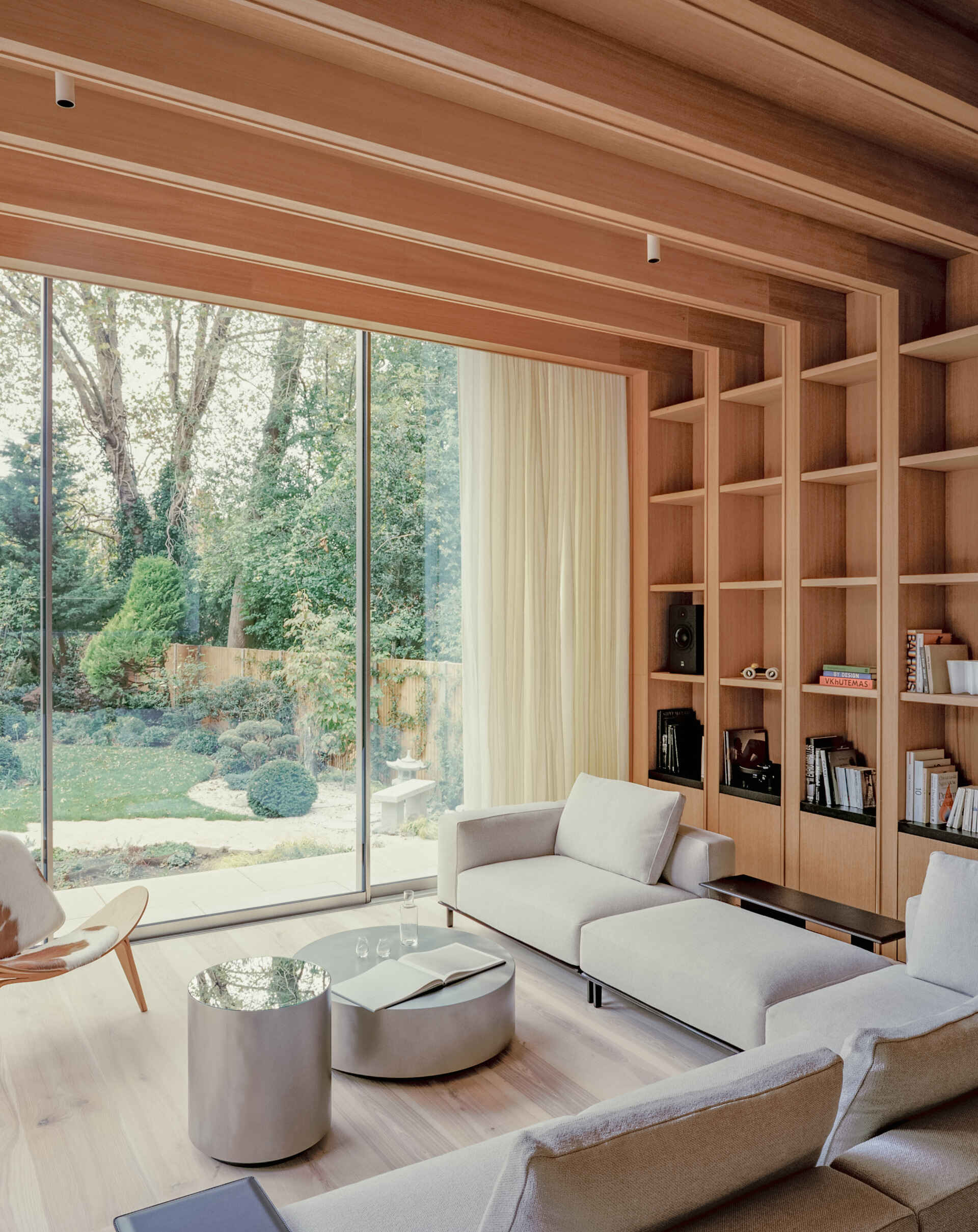 A sunlit living space steps down into the garden, using level changes and natural light to define the layout. The wood shelving in the living room adds a quieter layer to the space, balancing out the openness created by the glass walls.