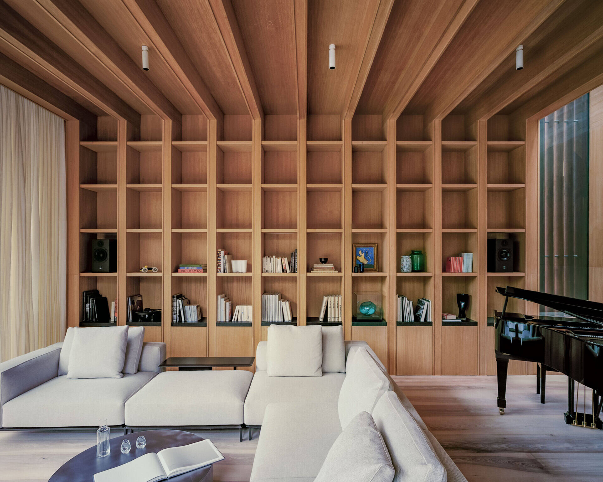 A sunlit living space steps down into the garden, using level changes and natural light to define the layout. The wood shelving in the living room adds a quieter layer to the space, balancing out the openness created by the glass walls.