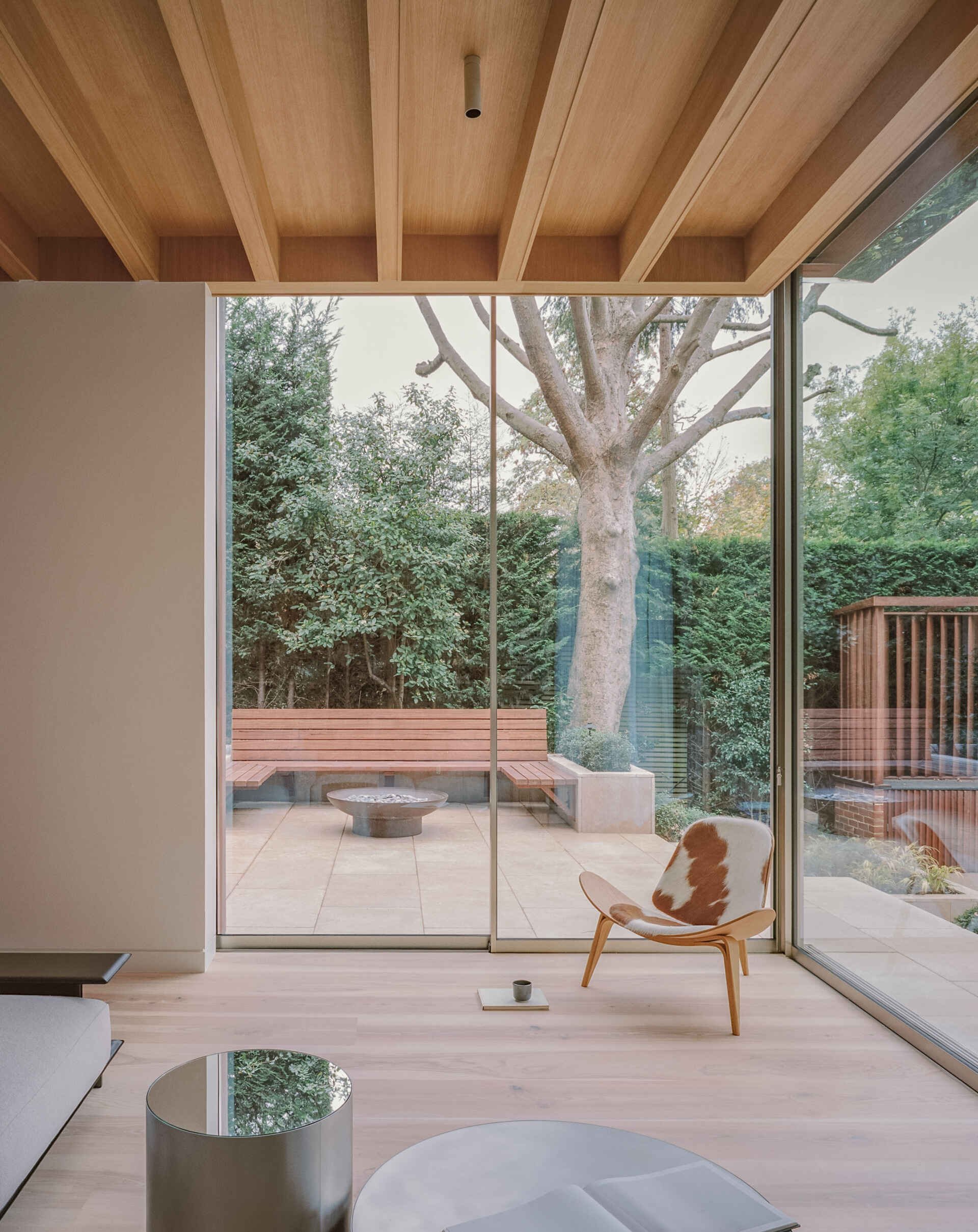 A sunlit living space steps down into the garden, using level changes and natural light to define the layout. The wood shelving in the living room adds a quieter layer to the space, balancing out the openness created by the glass walls.