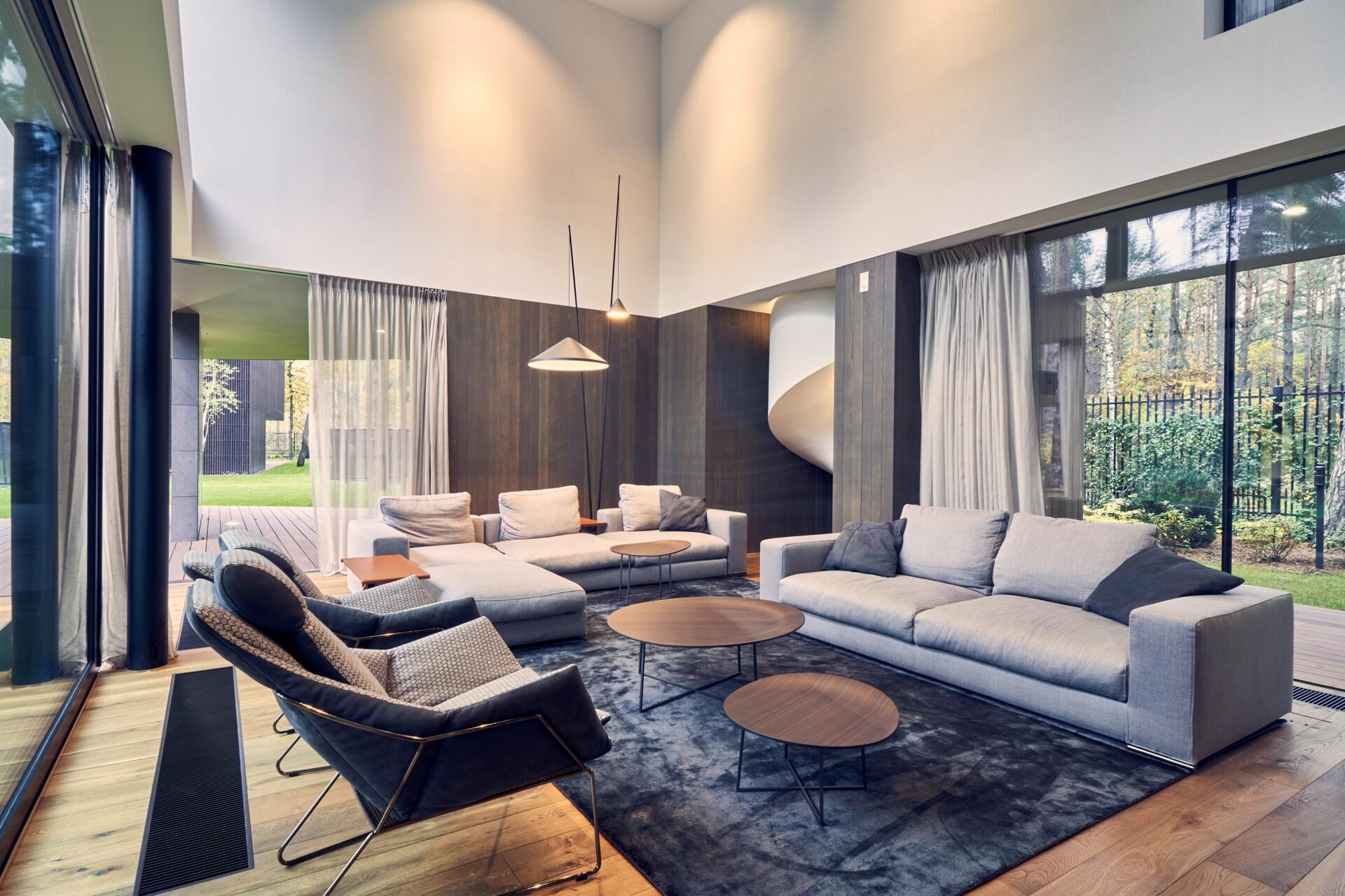 A double-height living room with warm wood floors and full-height windows creates a bright and open space connected to the outdoors.