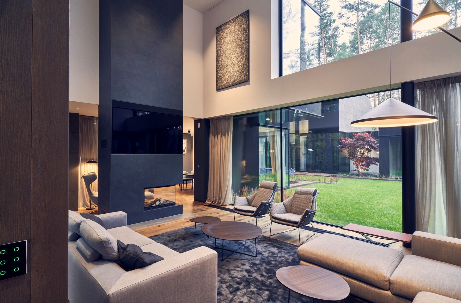 A double-height living room with warm wood floors and full-height windows creates a bright and open space connected to the outdoors.