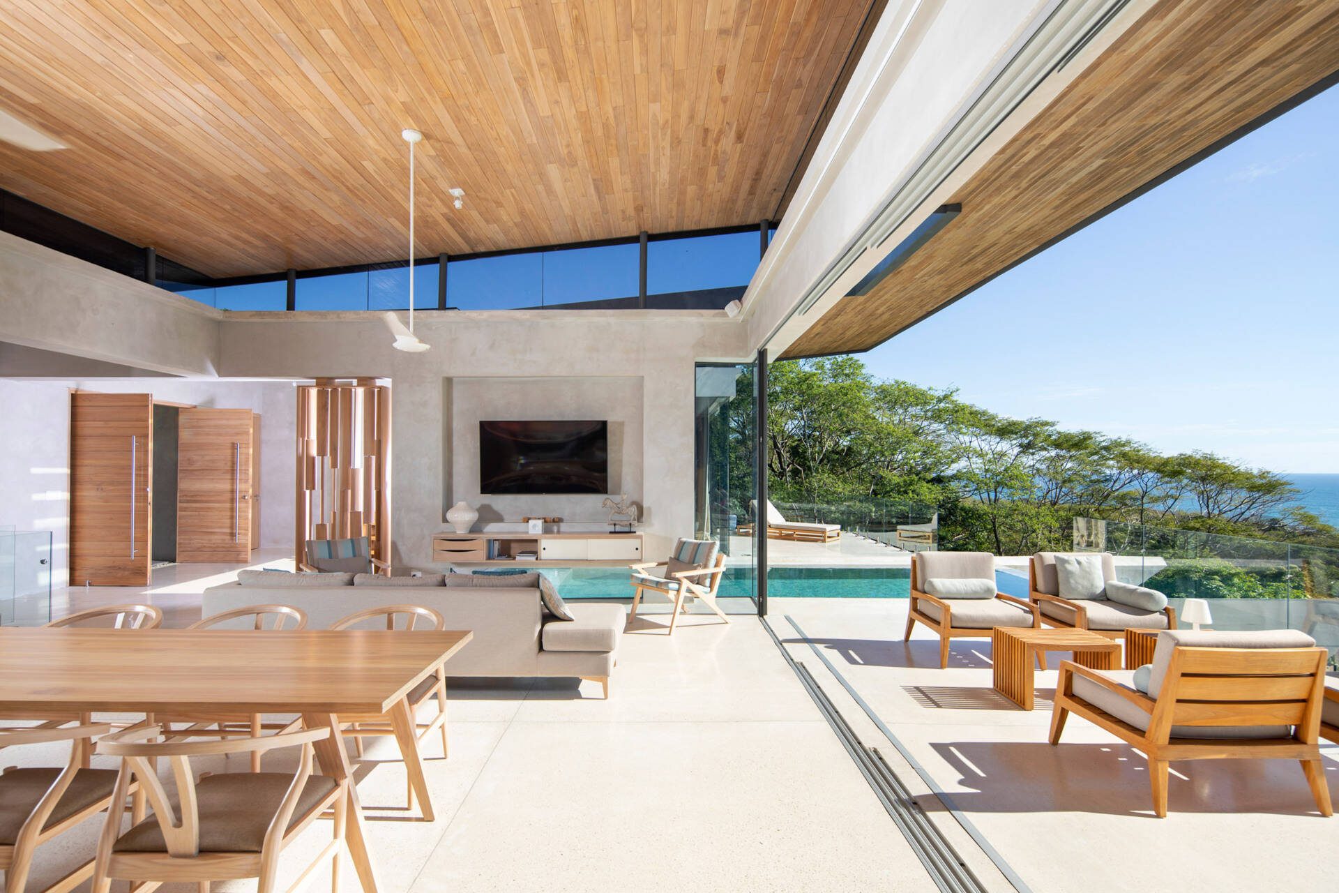 An open plan living space with a floating wood ceiling and sliding glass walls that open fully to a shaded balcony.