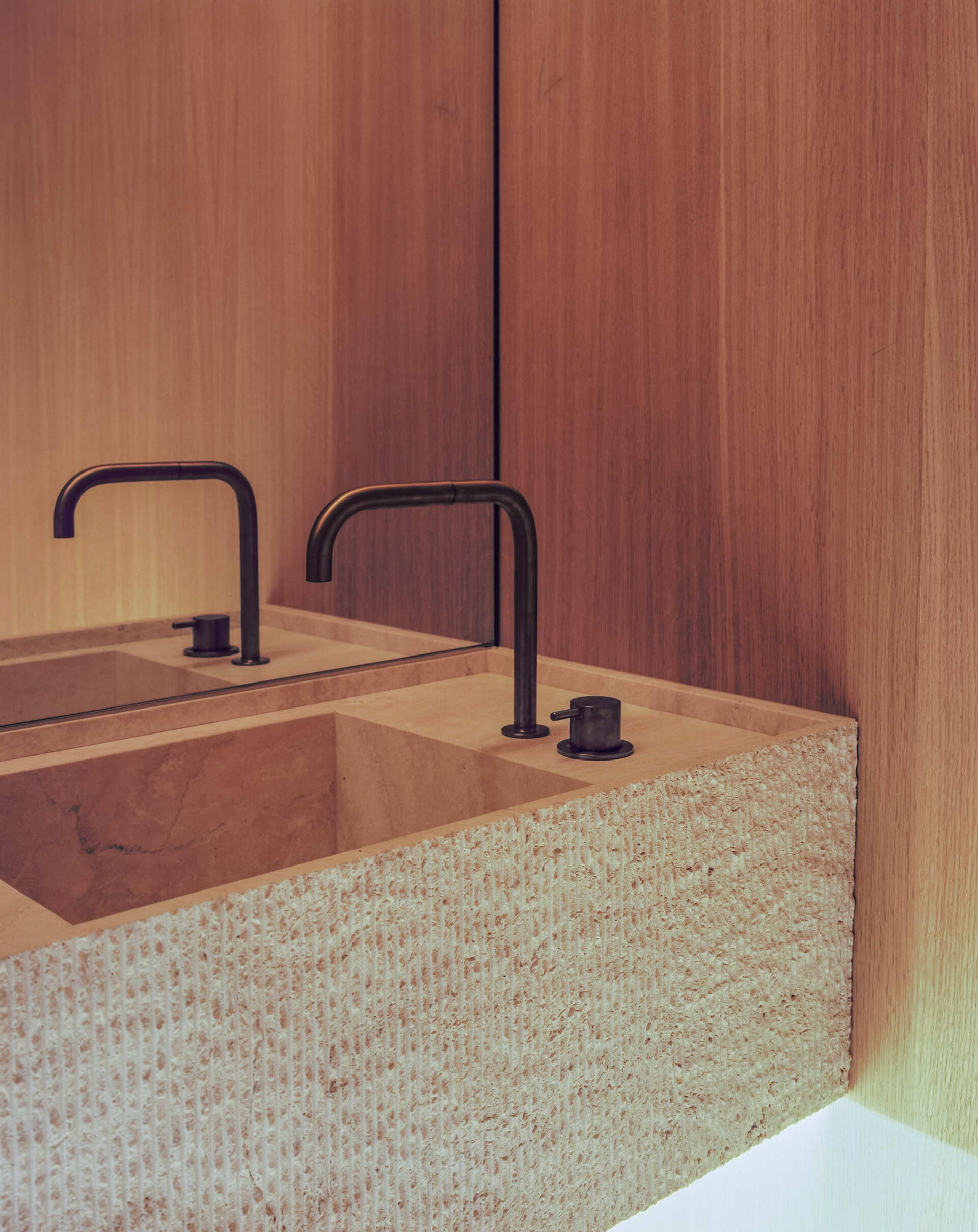 A powder room pairs raw texture with clean detailing, centered around a sculptural stone sink, its rough exterior contrasting with the smooth basin interior. Warm wood paneling wraps the space, while subtle underlighting lifts the heavy stone, giving it a floating effect and softening the overall look.