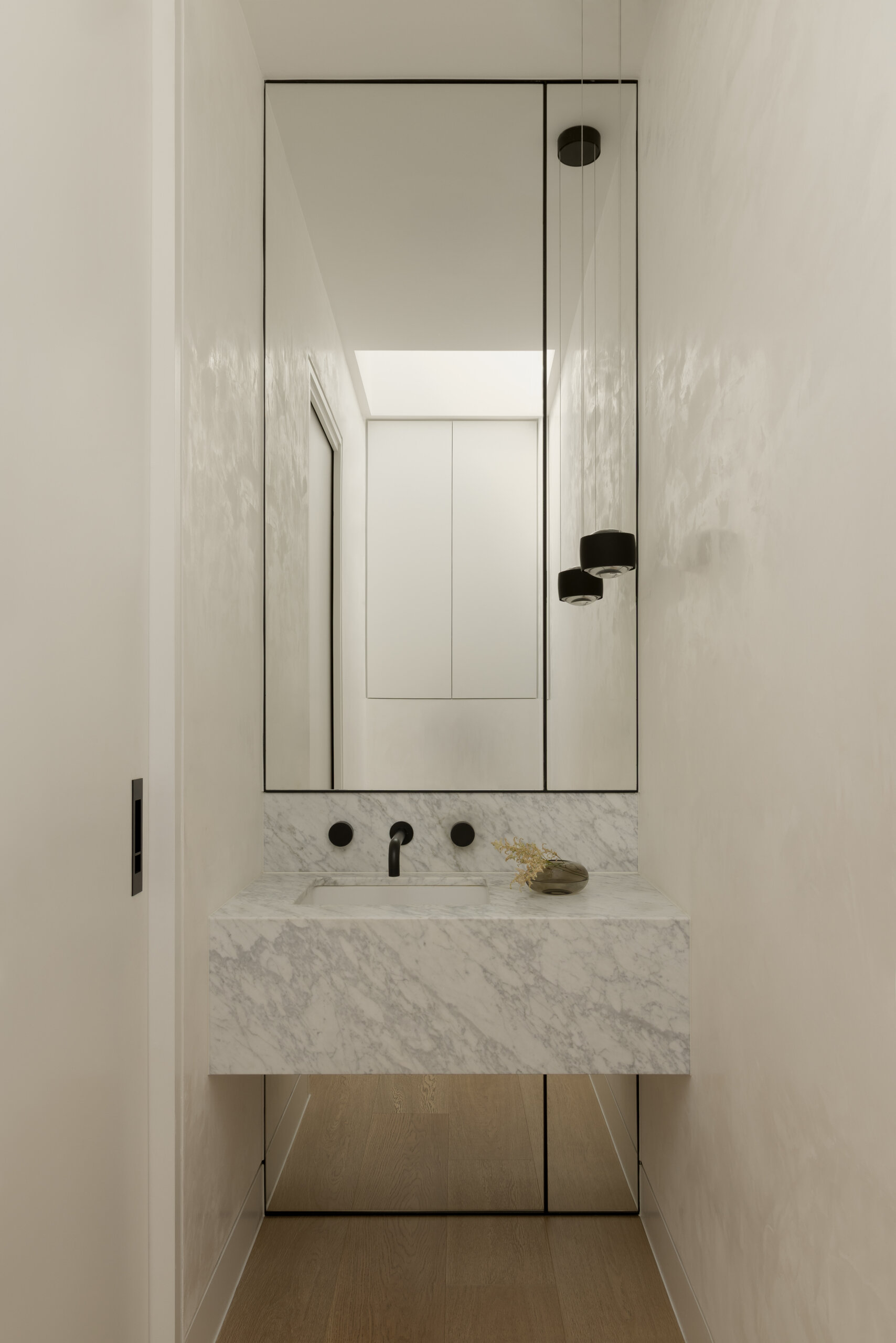 The powder room follows the same restrained material approach, offering a smaller but cohesive extension of the home’s overall design language. A tall mirror and white walls helps reflect light from the skylight, keeping the small space bright.