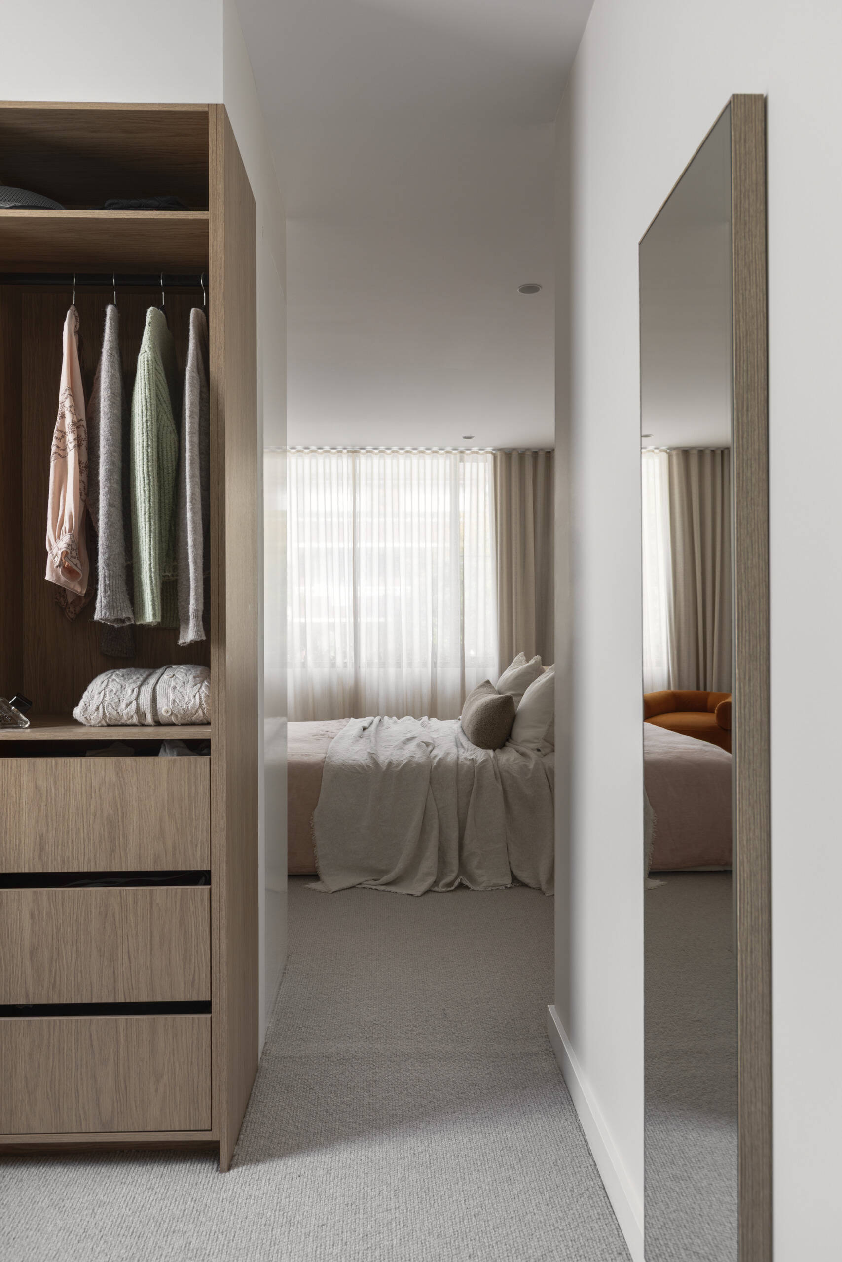Upstairs, this primary bedroom suite provides a more private zone for the homeowners. The bedroom has pale layered bedding, creamy walls, and full-height sheer curtains filtering the daylight into a gentle glow. The light wood wardrobe in its own area adds warmth and useful storage without crowding the room.
