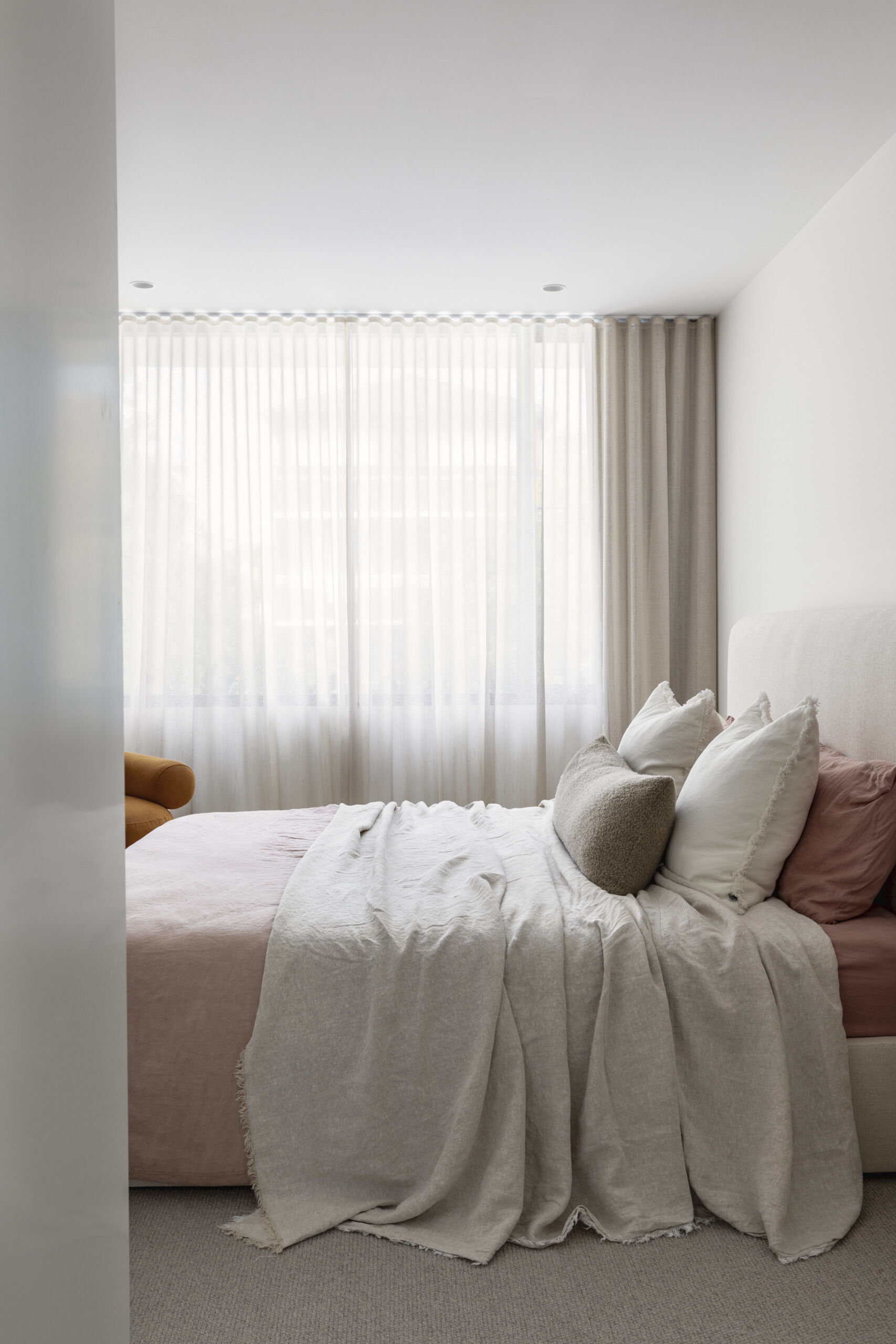 Upstairs, this primary bedroom suite provides a more private zone for the homeowners. The bedroom has pale layered bedding, creamy walls, and full-height sheer curtains filtering the daylight into a gentle glow. The light wood wardrobe in its own area adds warmth and useful storage without crowding the room.