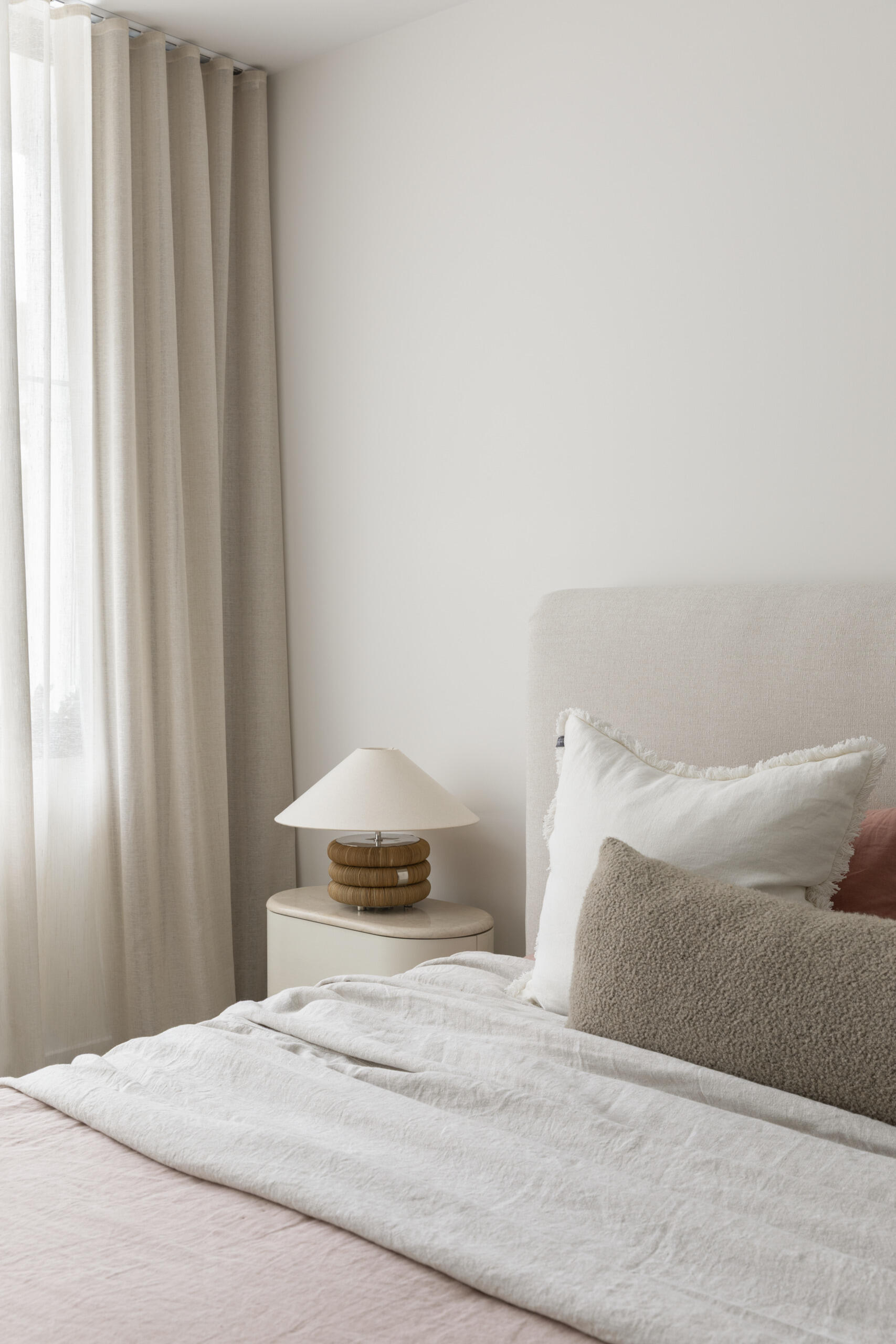 Upstairs, this primary bedroom suite provides a more private zone for the homeowners. The bedroom has pale layered bedding, creamy walls, and full-height sheer curtains filtering the daylight into a gentle glow. The light wood wardrobe in its own area adds warmth and useful storage without crowding the room.