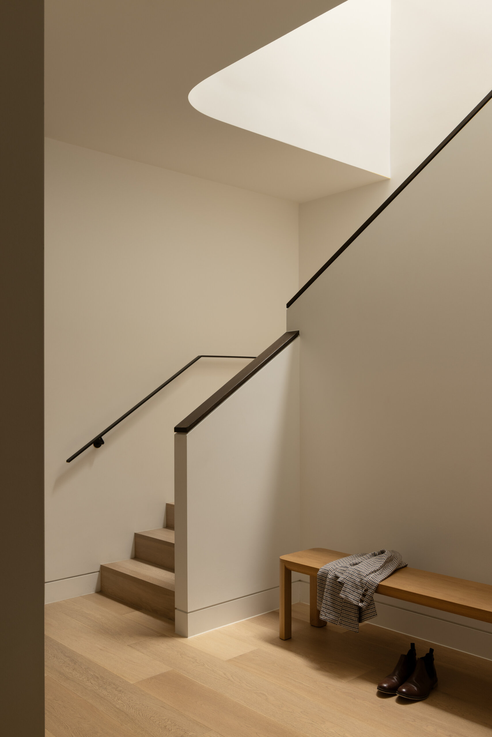 A softly lit staircase with a sculpted ceiling opening brings natural light into this minimal, warm transition from garage to home.