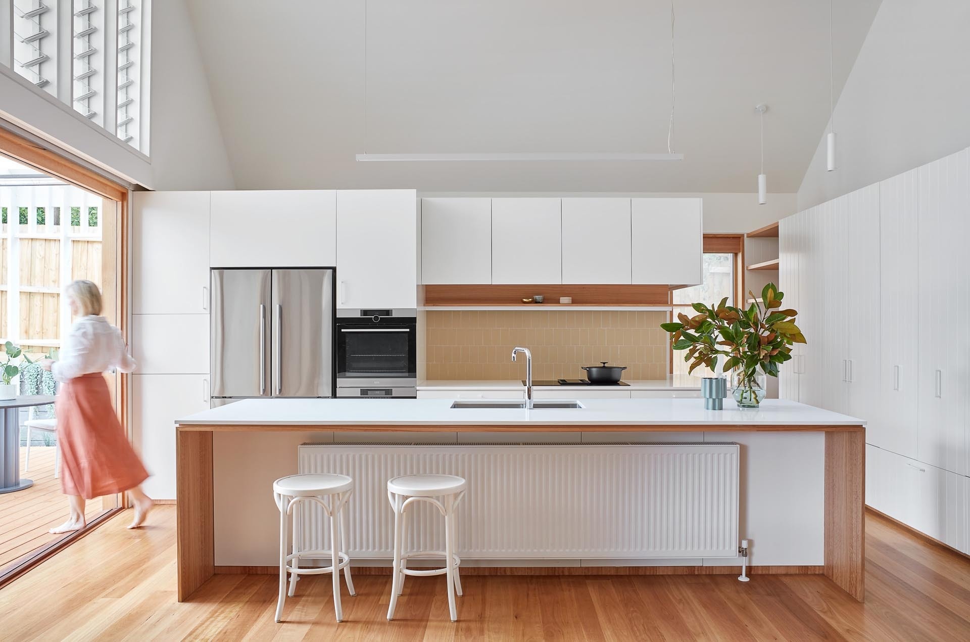 A clean, modern kitchen pairs white cabinetry with timber accents, opening directly to outdoor dining and supported by hidden functional spaces.