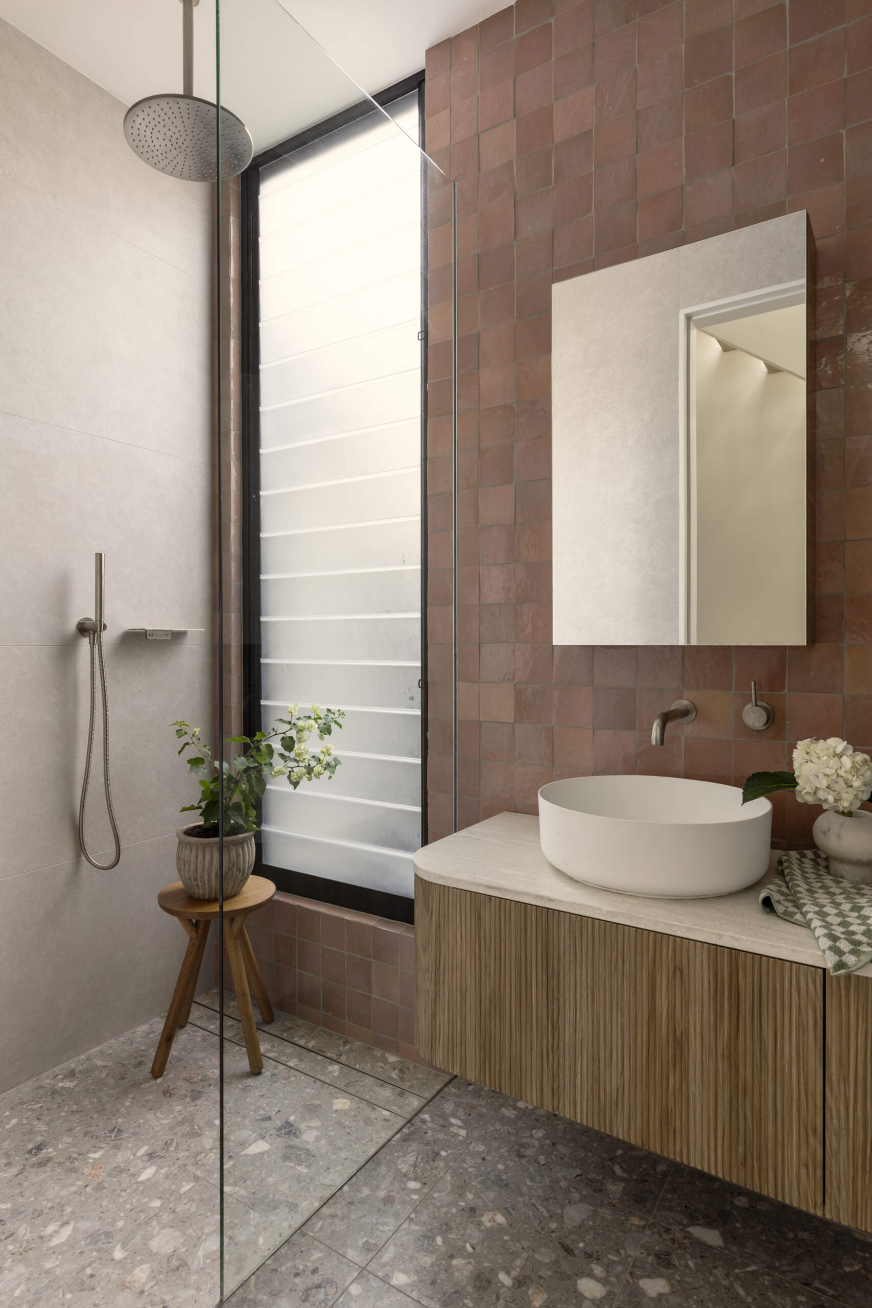 This guest bathroom sits nearby and pairs earthy terracotta-colored tile with soft gray surfaces for a look that feels warm and grounded. The ribbed wood vanity adds texture, while the round vessel sink keeps the lines clean and sculptural.
