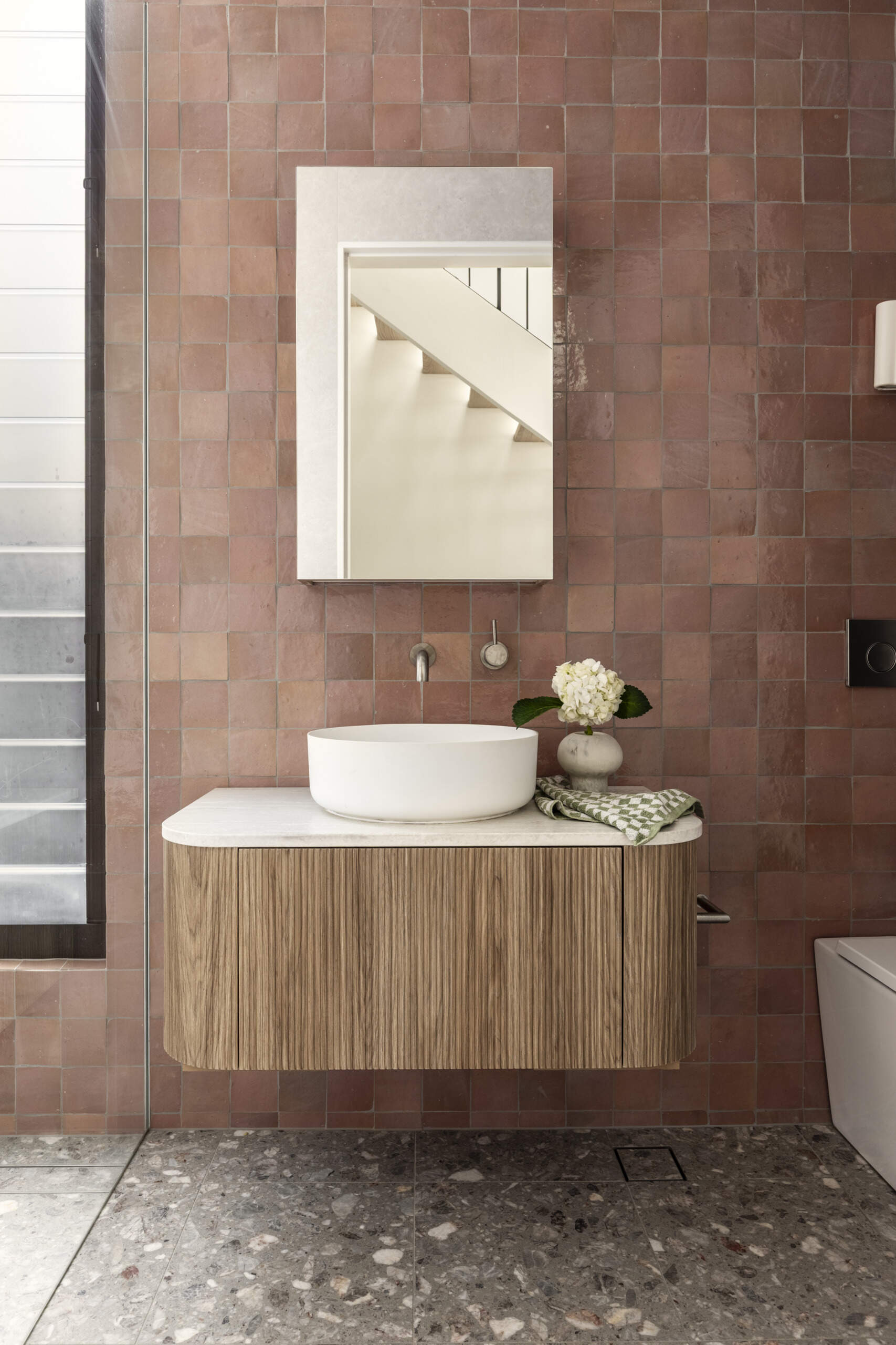 This guest bathroom sits nearby and pairs earthy terracotta-colored tile with soft gray surfaces for a look that feels warm and grounded. The ribbed wood vanity adds texture, while the round vessel sink keeps the lines clean and sculptural.