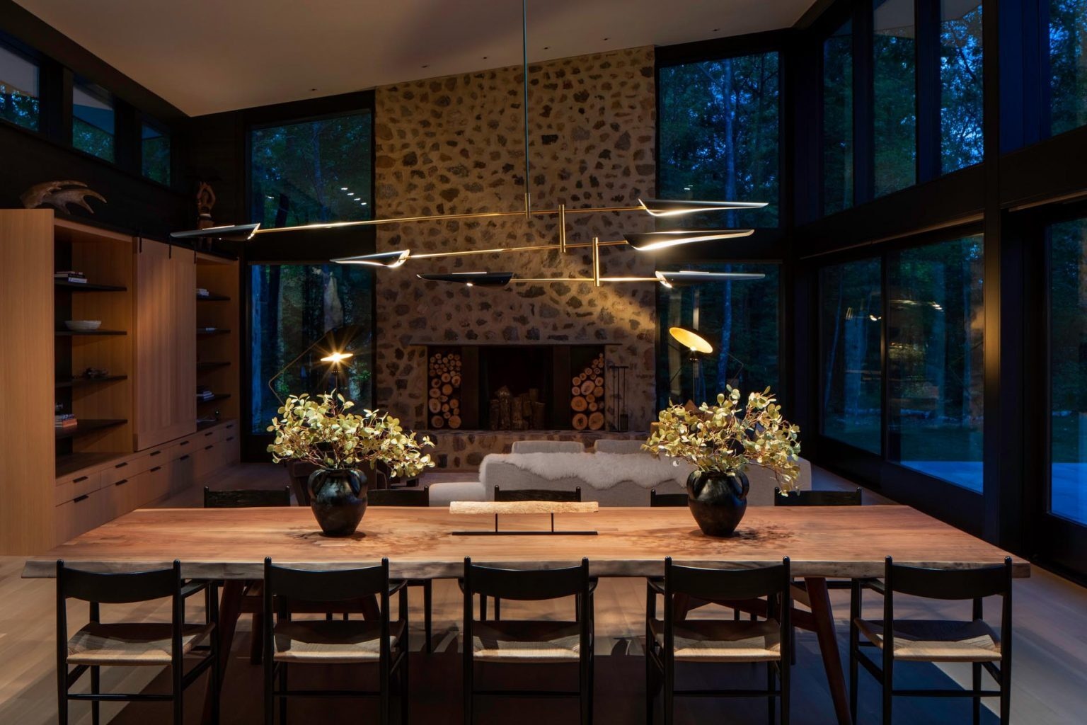 Una mesa de comedor de madera personalizada se combina con una lámpara de araña escultural y ofrece vistas a la espectacular chimenea de piedra.
