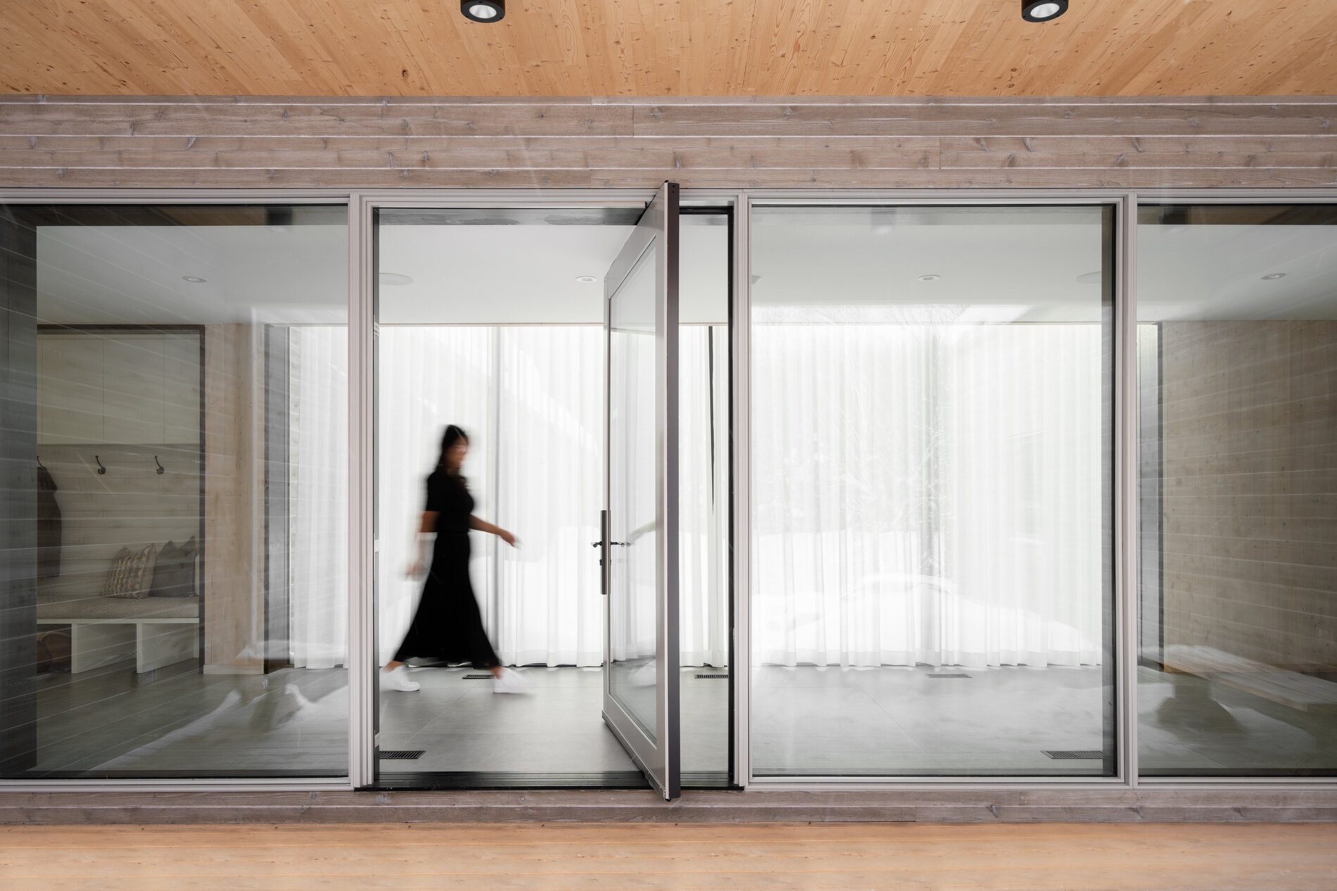 A glass-lined hallway that connects separate volumes, while also including a dedicated area for shoes, coats, storage, and benches.