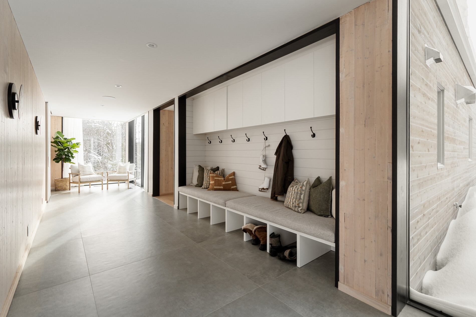 A glass-lined hallway that connects separate volumes, while also including a dedicated area for shoes, coats, storage, and benches.