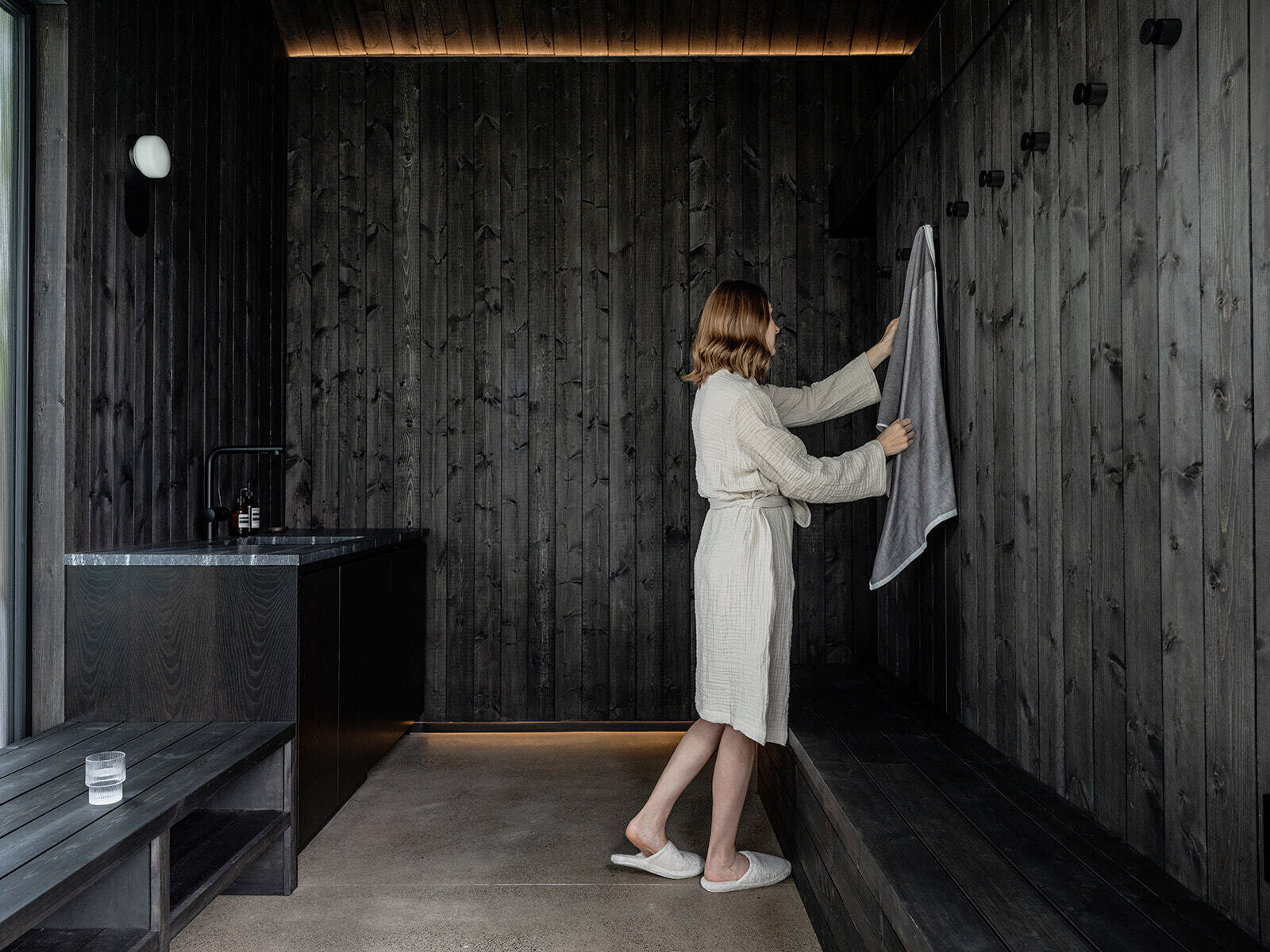 Inside Alameda Bathhouse, black-stained timber, textured stone, exposed concrete, oversized tiles, and soft hidden lighting create a warm and immersive atmosphere. Designed by Ment Architecture in Portland, Oregon, the spa-like interior features a sauna, steam room, and gathering space wrapped in rich materials and filtered natural light.