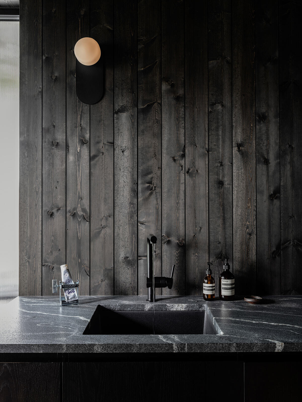 Inside Alameda Bathhouse, black-stained timber, textured stone, exposed concrete, oversized tiles, and soft hidden lighting create a warm and immersive atmosphere. Designed by Ment Architecture in Portland, Oregon, the spa-like interior features a sauna, steam room, and gathering space wrapped in rich materials and filtered natural light.