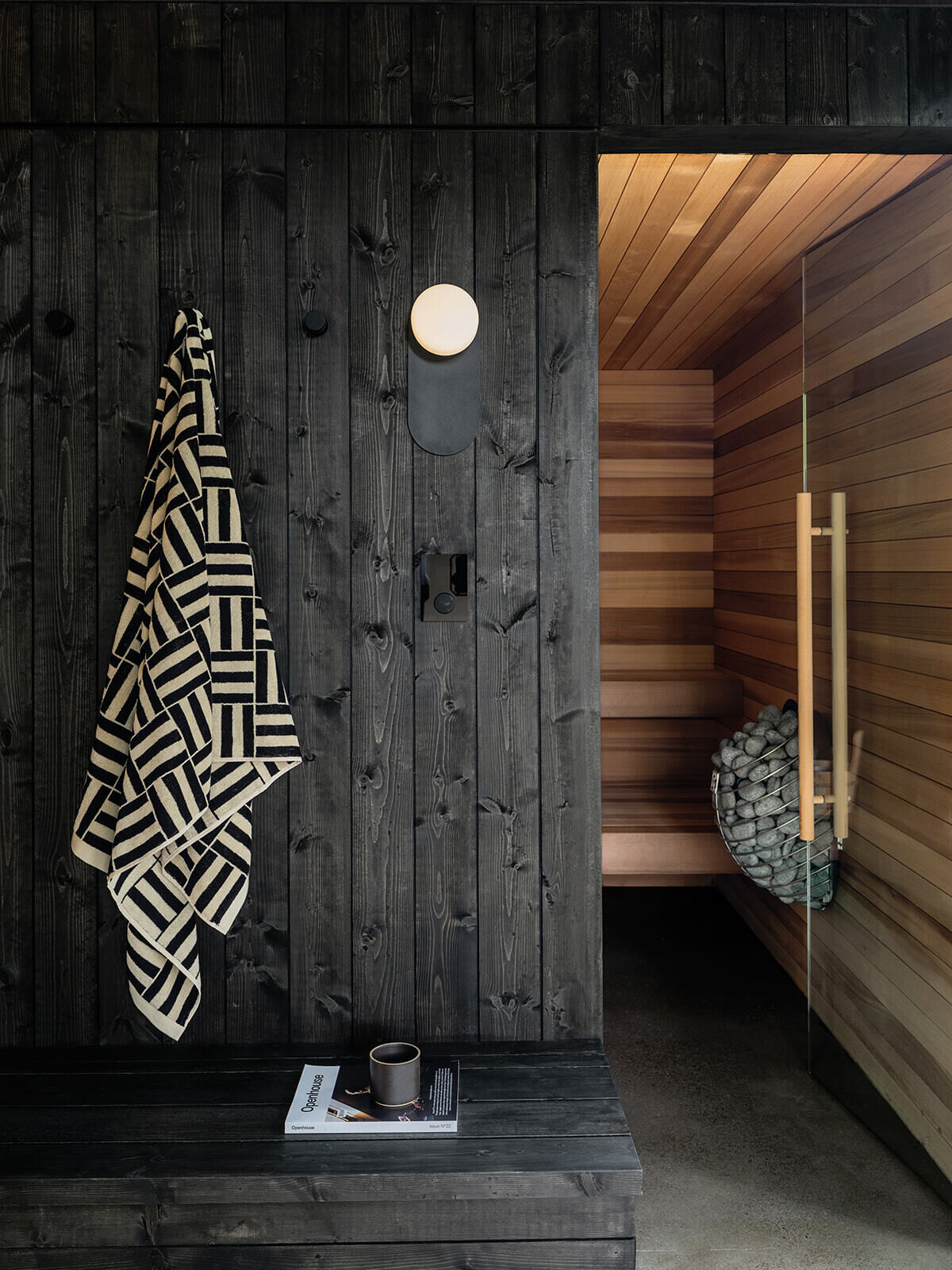 Inside Alameda Bathhouse, black-stained timber, textured stone, exposed concrete, oversized tiles, and soft hidden lighting create a warm and immersive atmosphere. Designed by Ment Architecture in Portland, Oregon, the spa-like interior features a sauna, steam room, and gathering space wrapped in rich materials and filtered natural light.