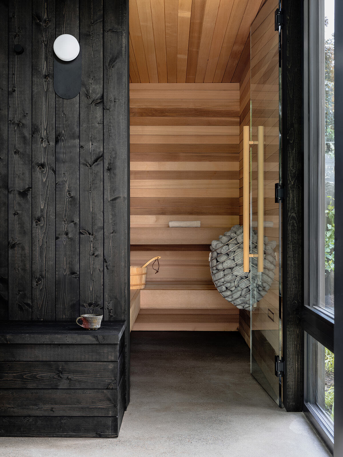 Inside Alameda Bathhouse, black-stained timber, textured stone, exposed concrete, oversized tiles, and soft hidden lighting create a warm and immersive atmosphere. Designed by Ment Architecture in Portland, Oregon, the spa-like interior features a sauna, steam room, and gathering space wrapped in rich materials and filtered natural light.