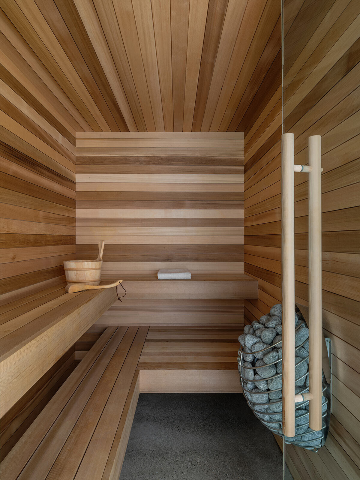 Inside Alameda Bathhouse, black-stained timber, textured stone, exposed concrete, oversized tiles, and soft hidden lighting create a warm and immersive atmosphere. Designed by Ment Architecture in Portland, Oregon, the spa-like interior features a sauna, steam room, and gathering space wrapped in rich materials and filtered natural light.