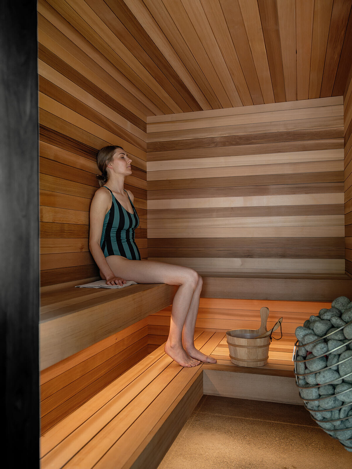 Inside Alameda Bathhouse, black-stained timber, textured stone, exposed concrete, oversized tiles, and soft hidden lighting create a warm and immersive atmosphere. Designed by Ment Architecture in Portland, Oregon, the spa-like interior features a sauna, steam room, and gathering space wrapped in rich materials and filtered natural light.