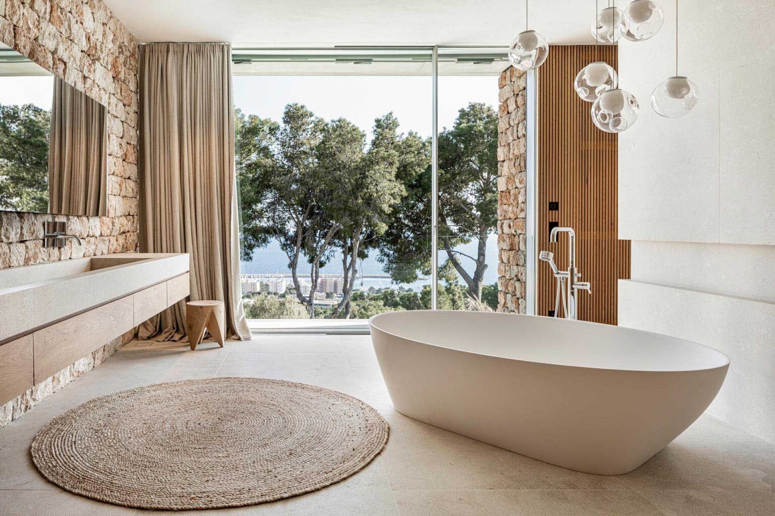 This Mallorca bathroom pairs textured stone walls with a floating vanity and a freestanding bathtub overlooking the sea.