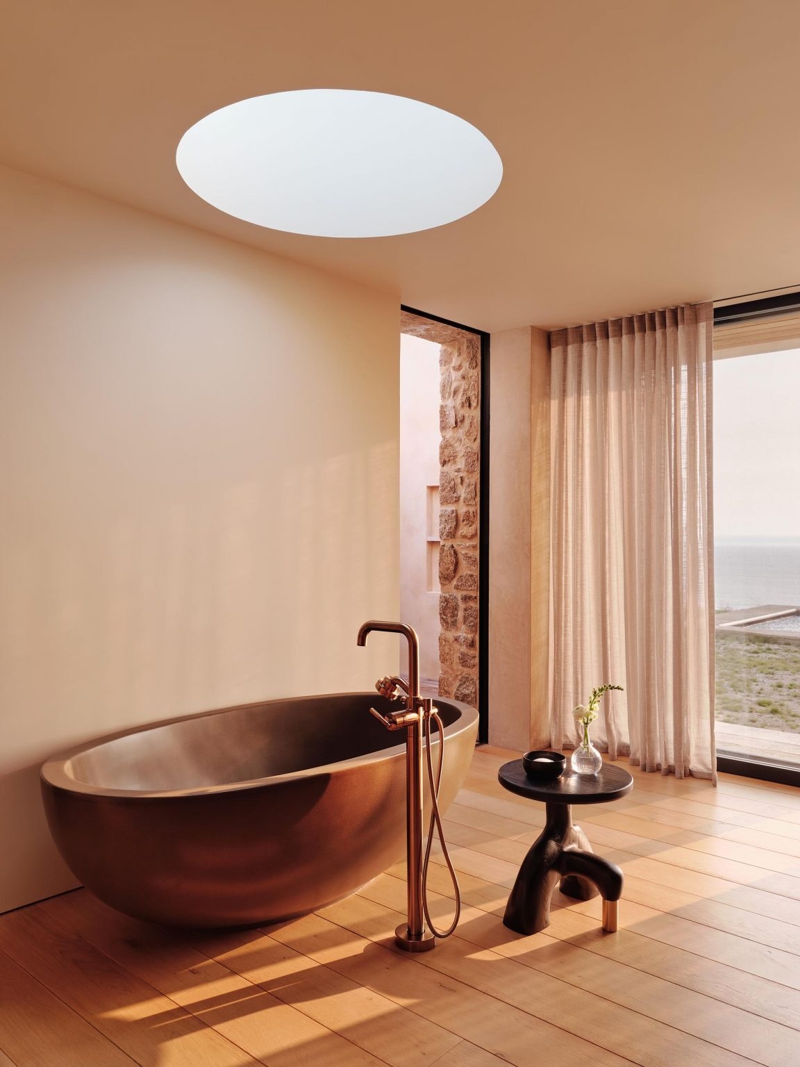 In the en-suite bathroom, a freestanding bathtub sits beneath a circular skylight that draws natural light into the space from above. Wood flooring softens the room and adds warmth against the stone and plaster finishes.