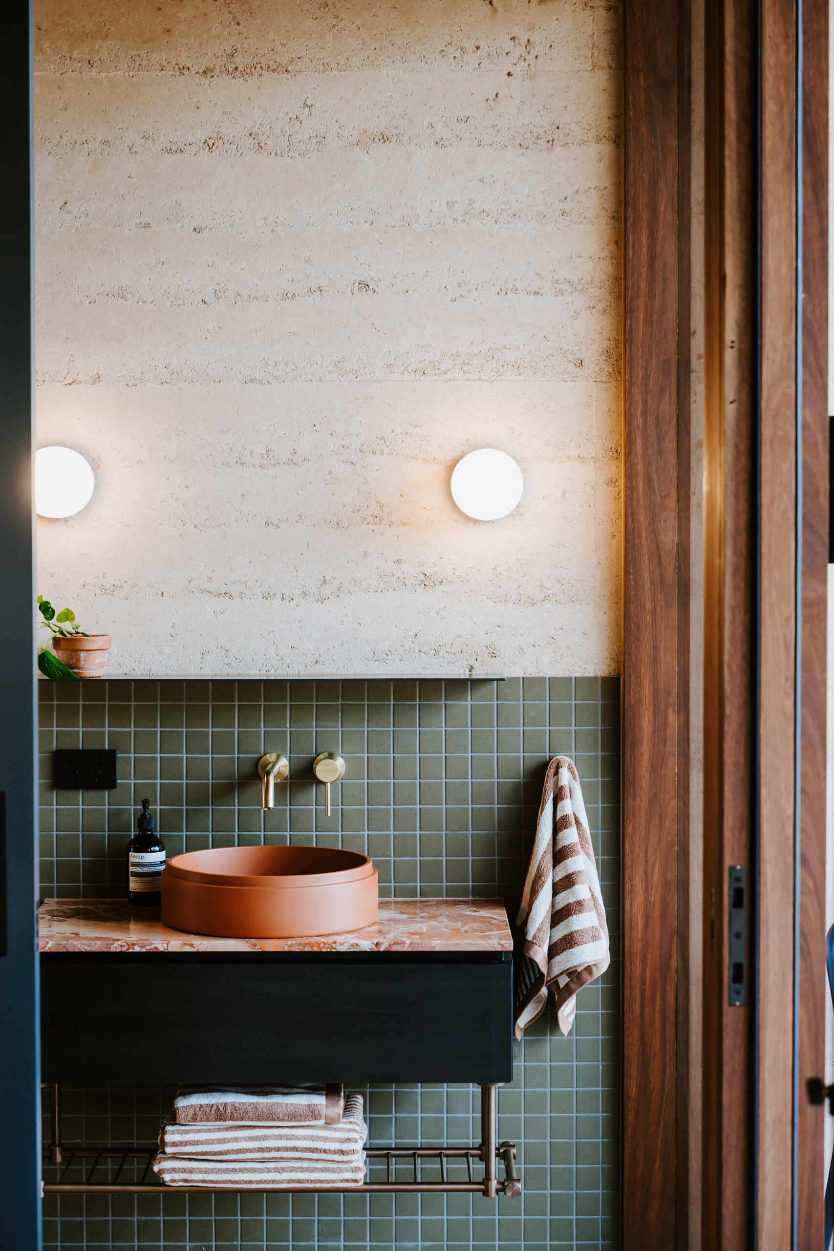 A modern off-grid cabin with rammed earth walls and sage green square tiles in the bathroom.