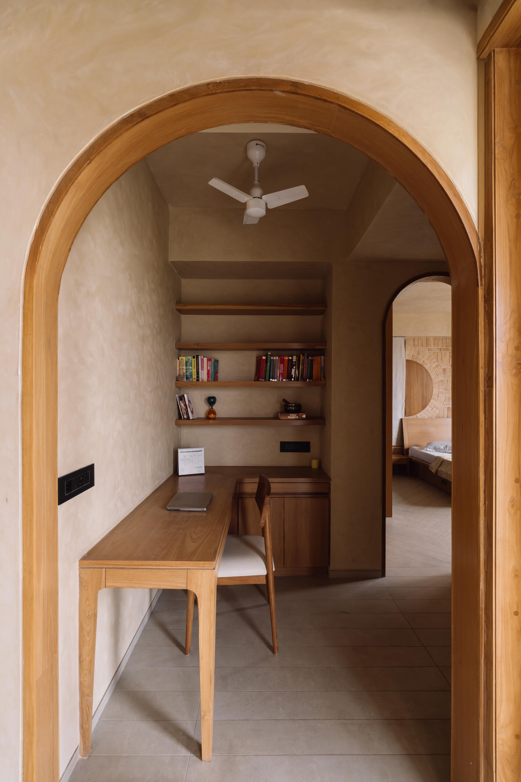 This warm minimalist study nook features floating timber shelves, a built-in desk, soft neutral tones, and an arched doorway inside an Ahmedabad apartment by UA Lab.