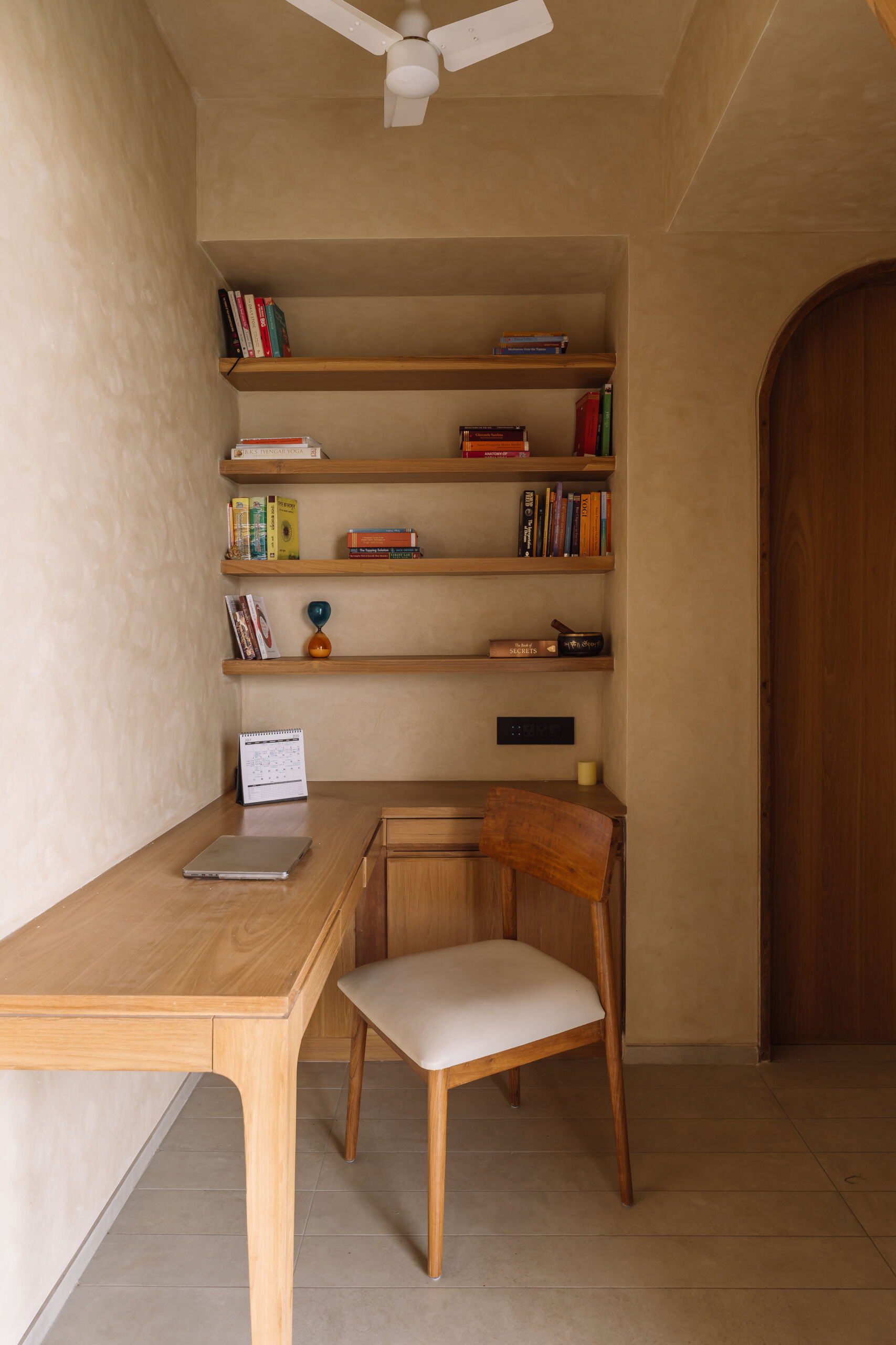 This warm minimalist study nook features floating timber shelves, a built-in desk, soft neutral tones, and an arched doorway inside an Ahmedabad apartment by UA Lab.