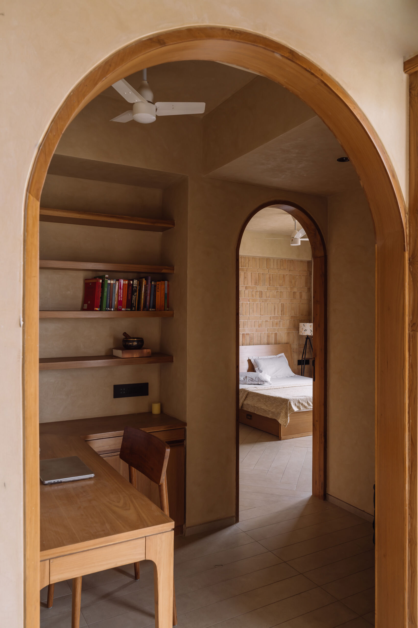 This warm minimalist study nook features floating timber shelves, a built-in desk, soft neutral tones, and an arched doorway inside an Ahmedabad apartment by UA Lab.