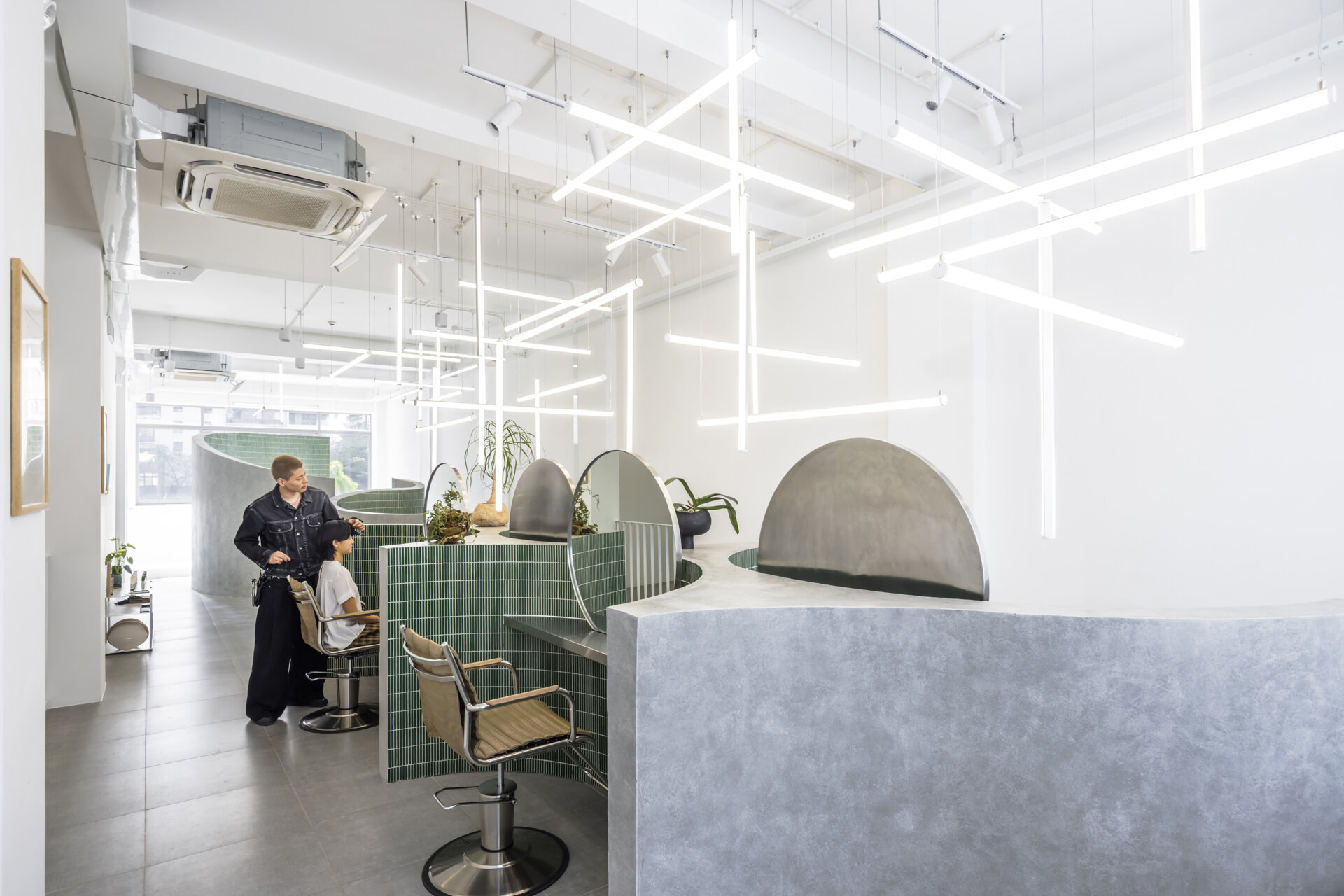 A hair salon in Ho Chi Minh City designed by Inrestudio, shaped by a continuous sculptural wall that guides movement through waiting areas, styling stations, and shampoo spaces. The interior shifts between green tile and raw grey surfaces, with light and form changing as you move through each level of the maisonette.