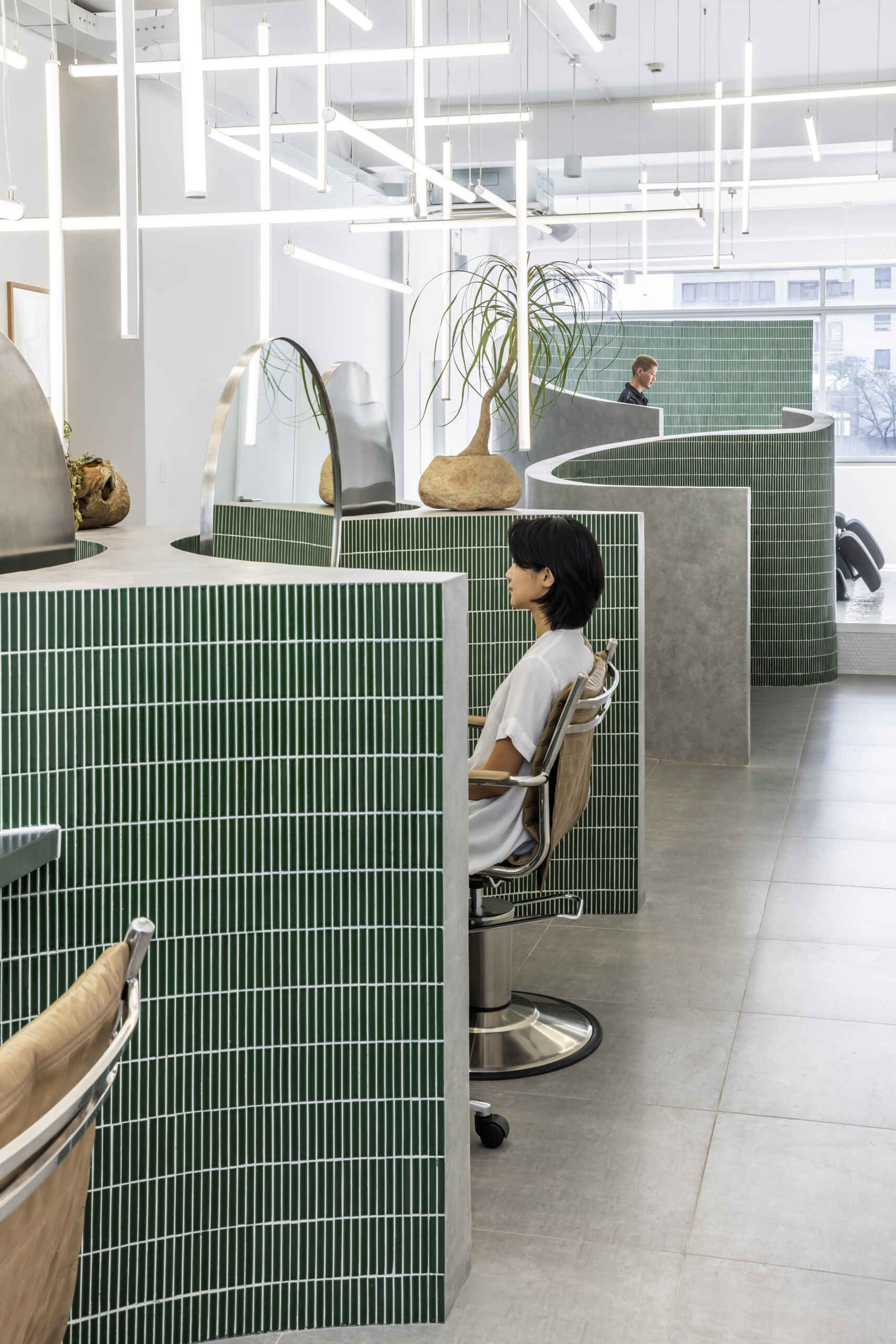 A hair salon in Ho Chi Minh City designed by Inrestudio, shaped by a continuous sculptural wall that guides movement through waiting areas, styling stations, and shampoo spaces. The interior shifts between green tile and raw grey surfaces, with light and form changing as you move through each level of the maisonette.