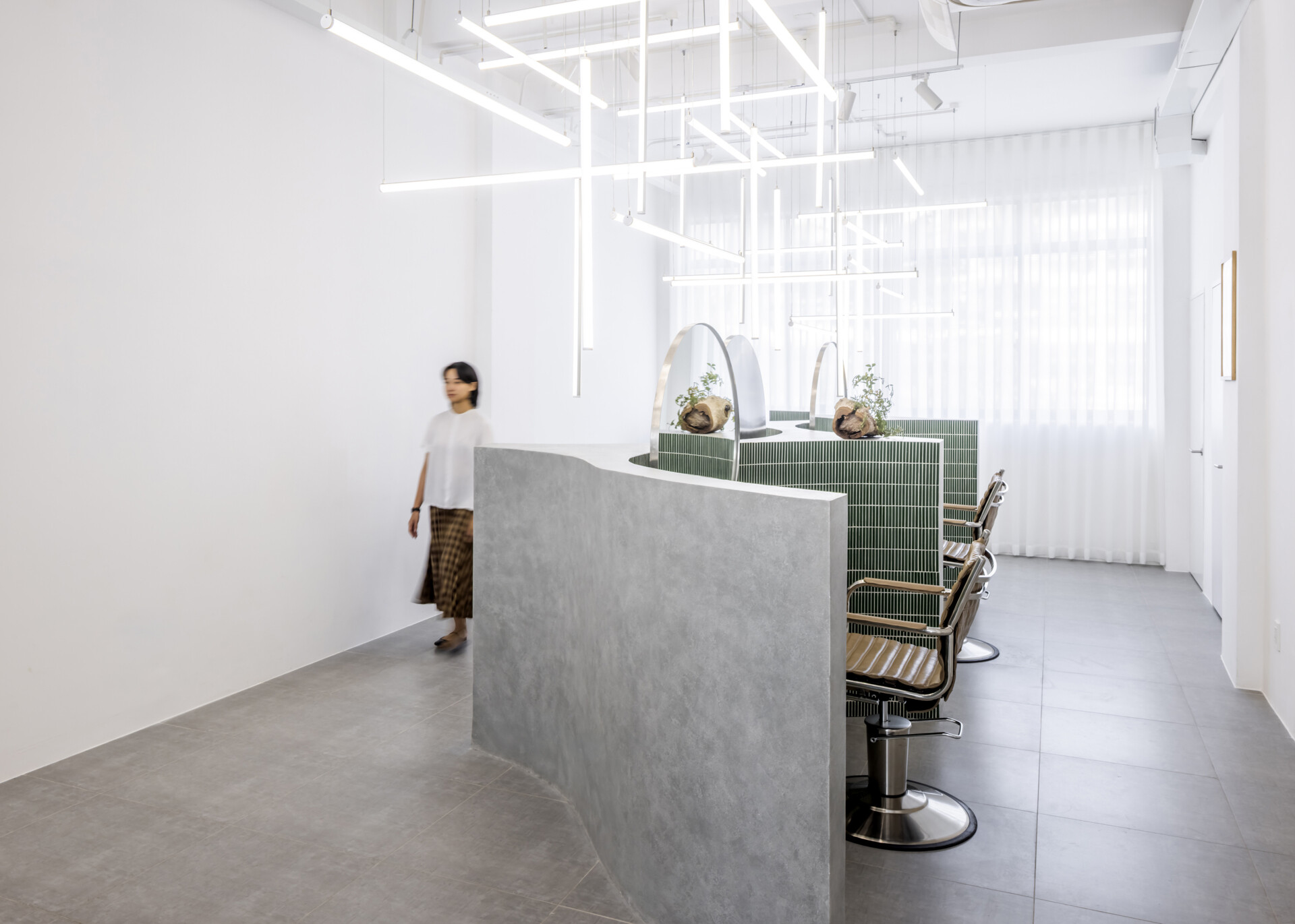 A hair salon in Ho Chi Minh City designed by Inrestudio, shaped by a continuous sculptural wall that guides movement through waiting areas, styling stations, and shampoo spaces. The interior shifts between green tile and raw grey surfaces, with light and form changing as you move through each level of the maisonette.