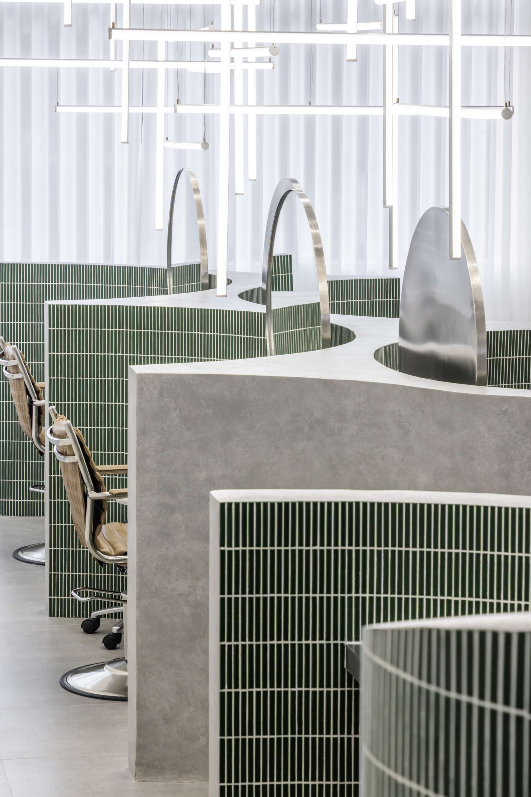 A hair salon in Ho Chi Minh City designed by Inrestudio, shaped by a continuous sculptural wall that guides movement through waiting areas, styling stations, and shampoo spaces. The interior shifts between green tile and raw grey surfaces, with light and form changing as you move through each level of the maisonette.
