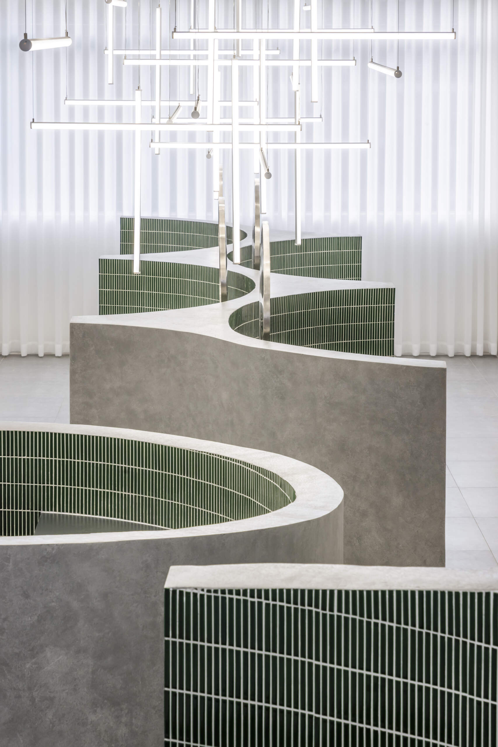 A hair salon in Ho Chi Minh City designed by Inrestudio, shaped by a continuous sculptural wall that guides movement through waiting areas, styling stations, and shampoo spaces. The interior shifts between green tile and raw grey surfaces, with light and form changing as you move through each level of the maisonette.