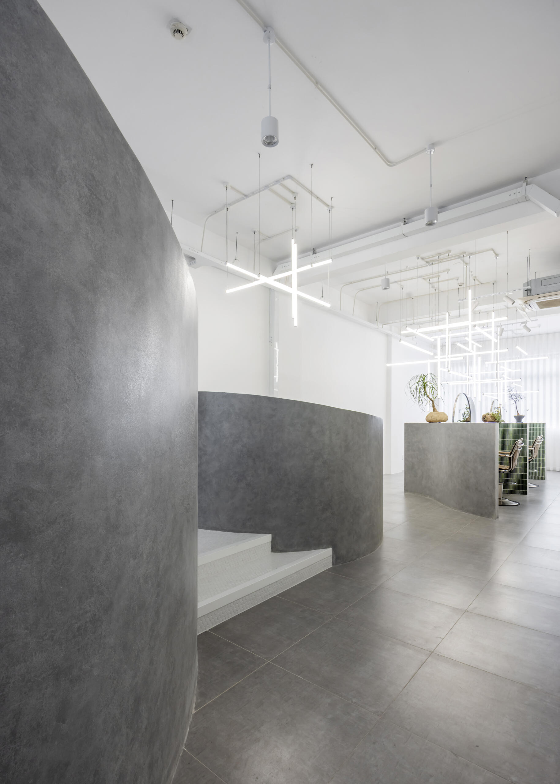 A hair salon in Ho Chi Minh City designed by Inrestudio, shaped by a continuous sculptural wall that guides movement through waiting areas, styling stations, and shampoo spaces. The interior shifts between green tile and raw grey surfaces, with light and form changing as you move through each level of the maisonette.