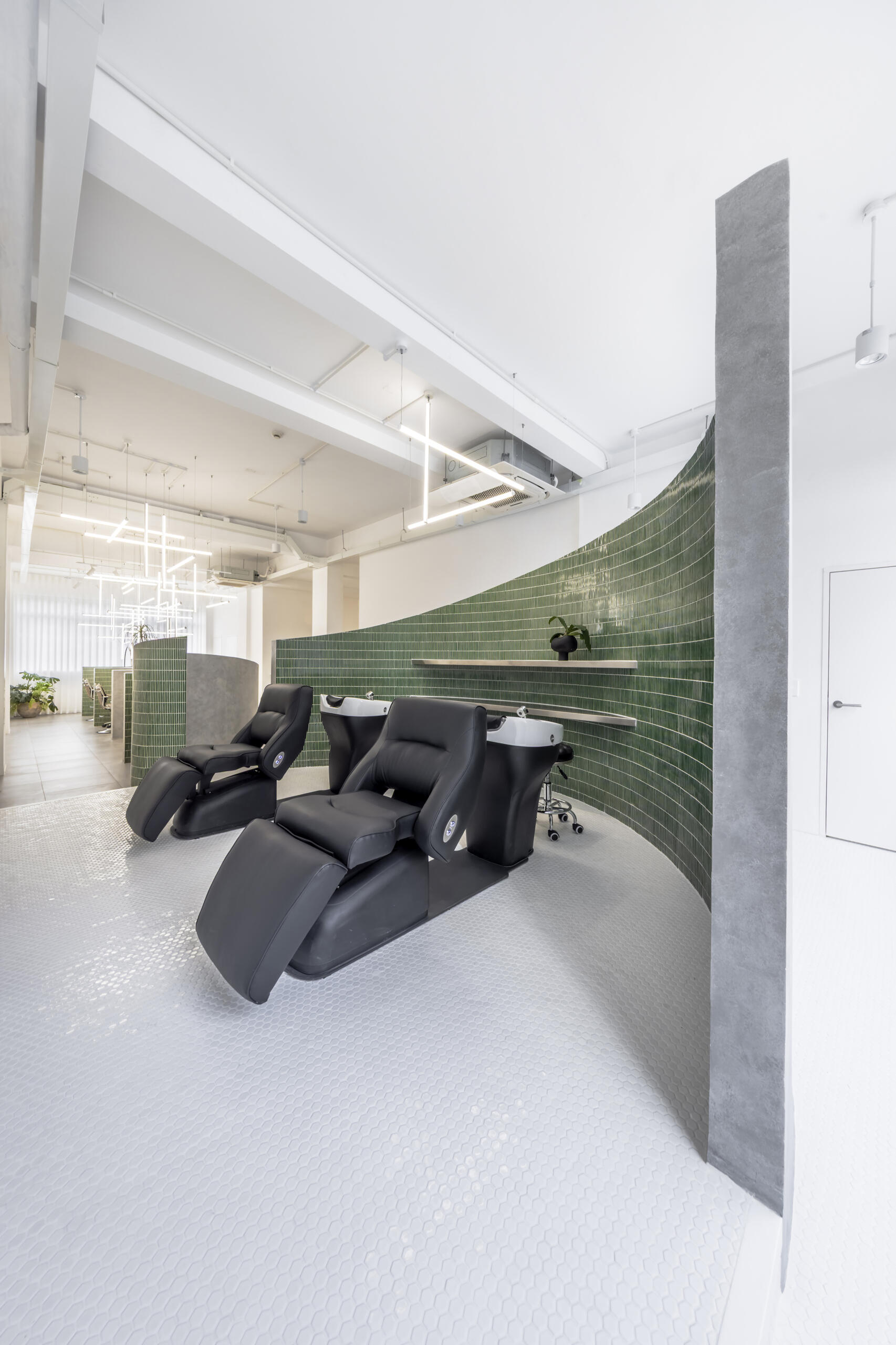 A hair salon in Ho Chi Minh City designed by Inrestudio, shaped by a continuous sculptural wall that guides movement through waiting areas, styling stations, and shampoo spaces. The interior shifts between green tile and raw grey surfaces, with light and form changing as you move through each level of the maisonette.