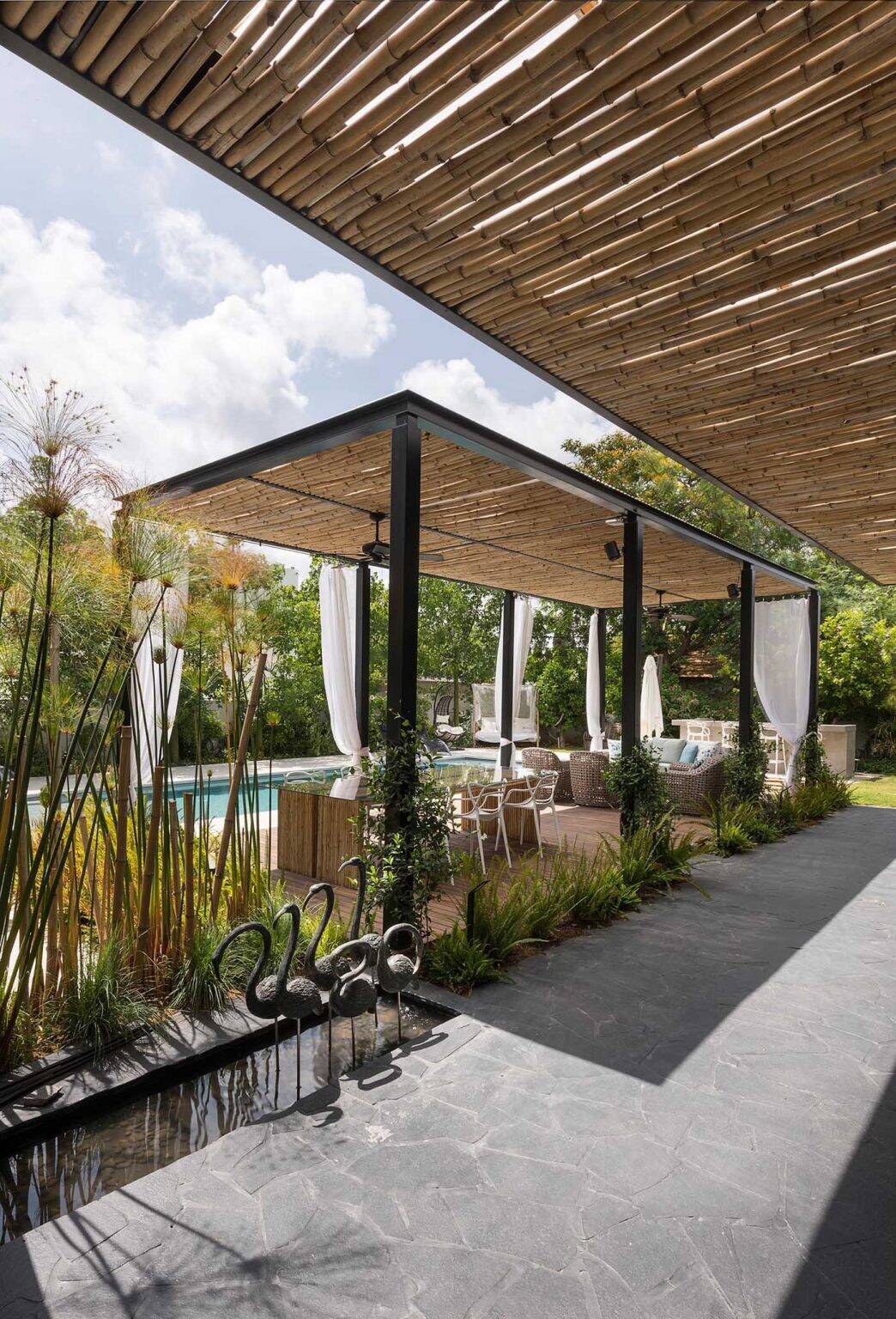 A bamboo pergola with white curtains creates a stylish poolside outdoor living and dining area beside this modern home.