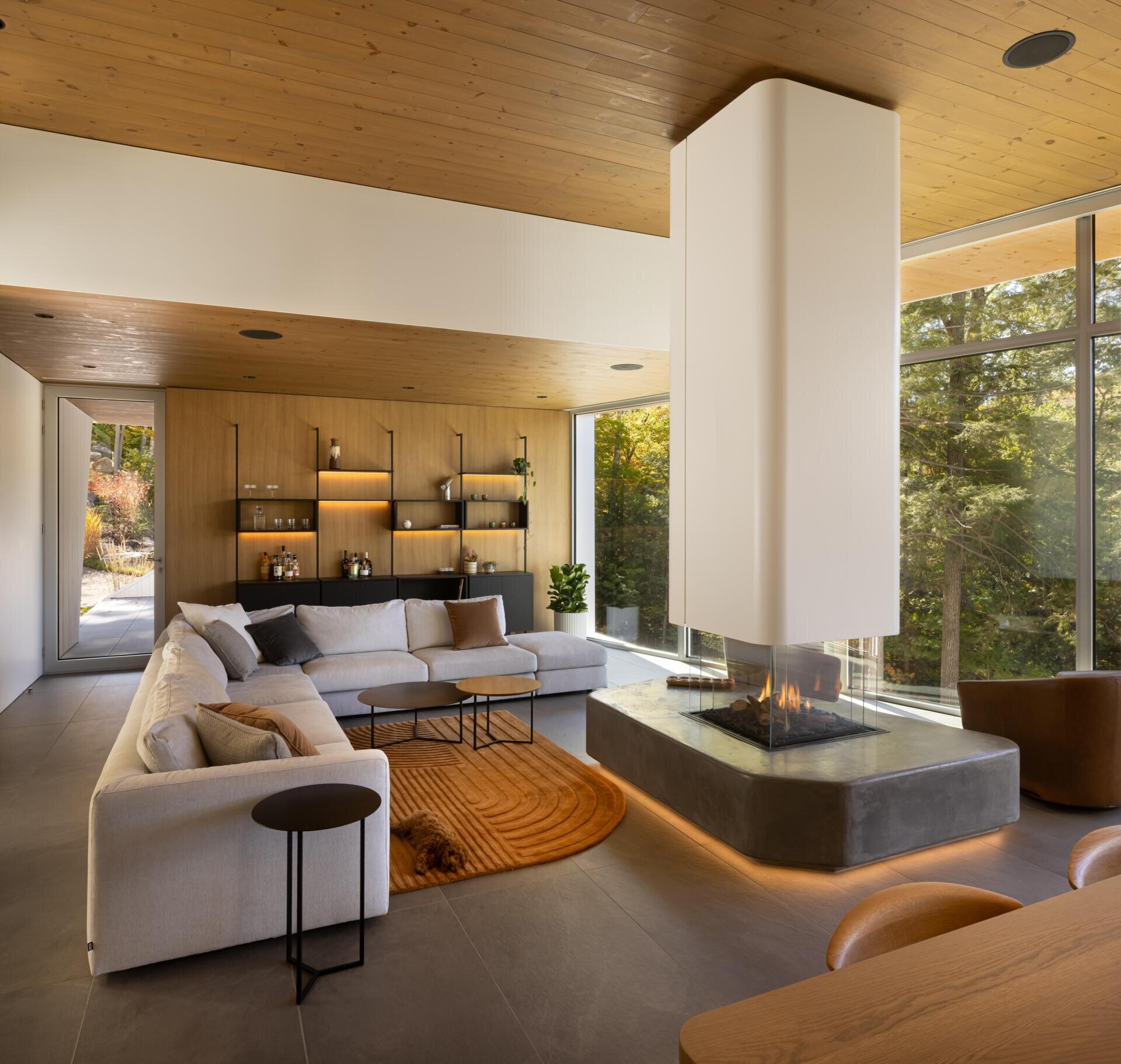 This lakeside living room combines soaring views, warm timber finishes, and a sculptural fireplace inside a contemporary Québec home.