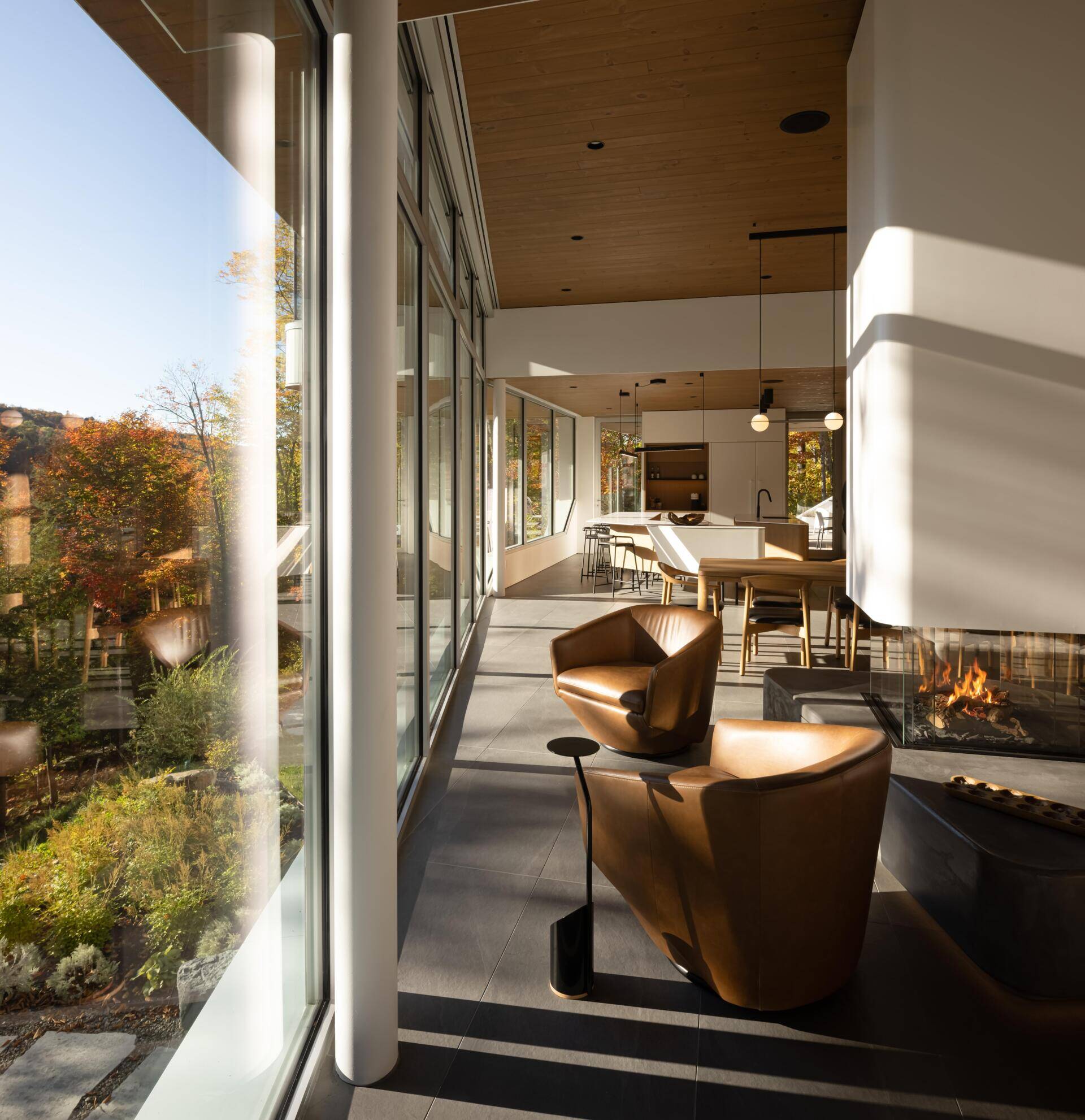 This lakeside living room combines soaring views, warm timber finishes, and a sculptural fireplace inside a contemporary Québec home.