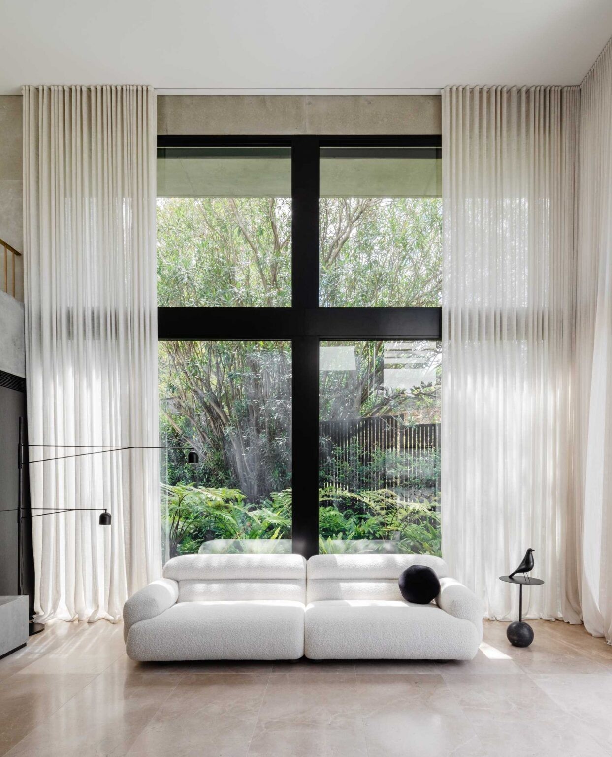 The double-height living room in this Sydney home combines concrete walls, limestone floors, skylights, and full-height glazing with views of lush gardens and palms.
