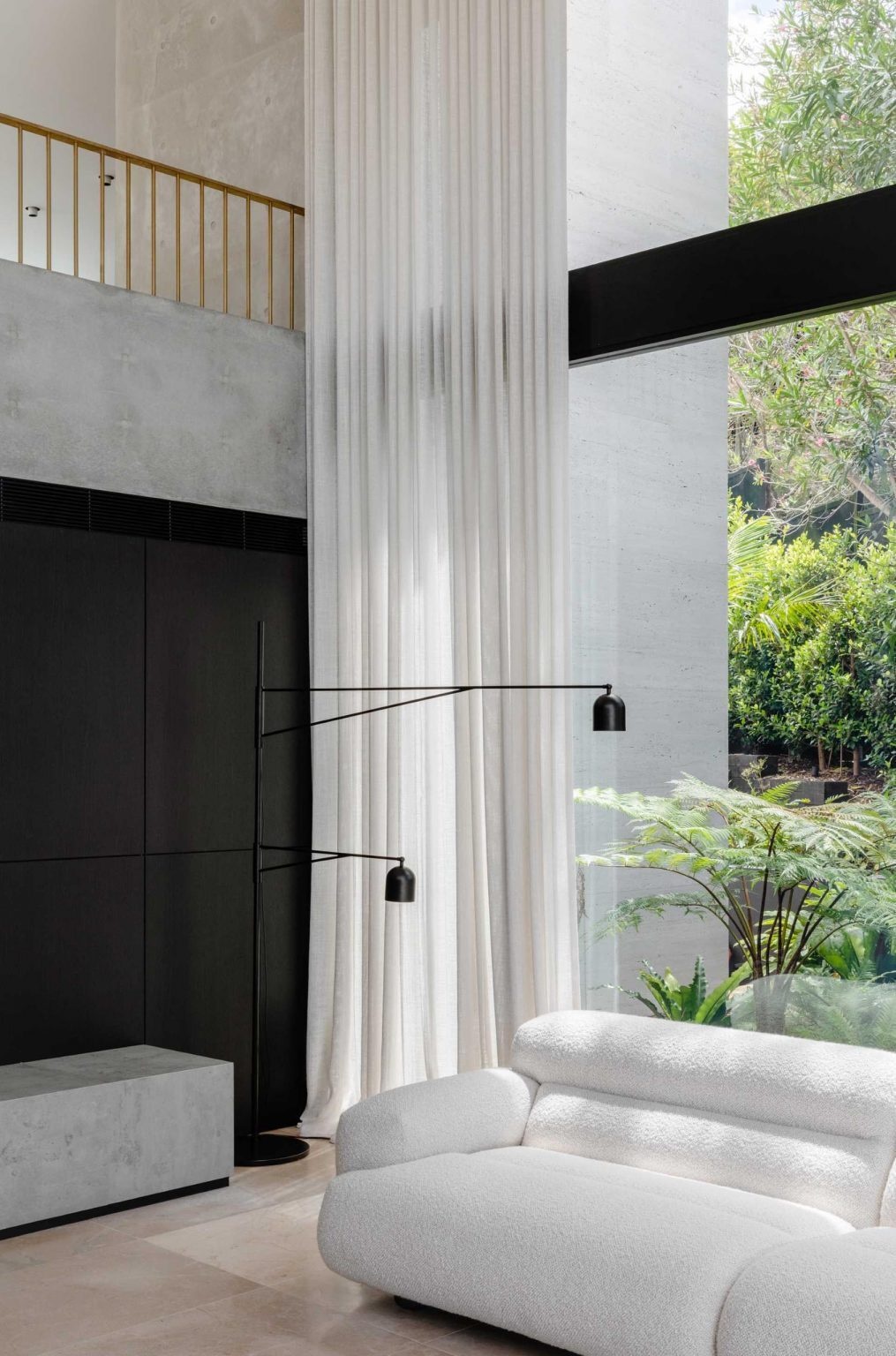 The double-height living room in this Sydney home combines concrete walls, limestone floors, skylights, and full-height glazing with views of lush gardens and palms.