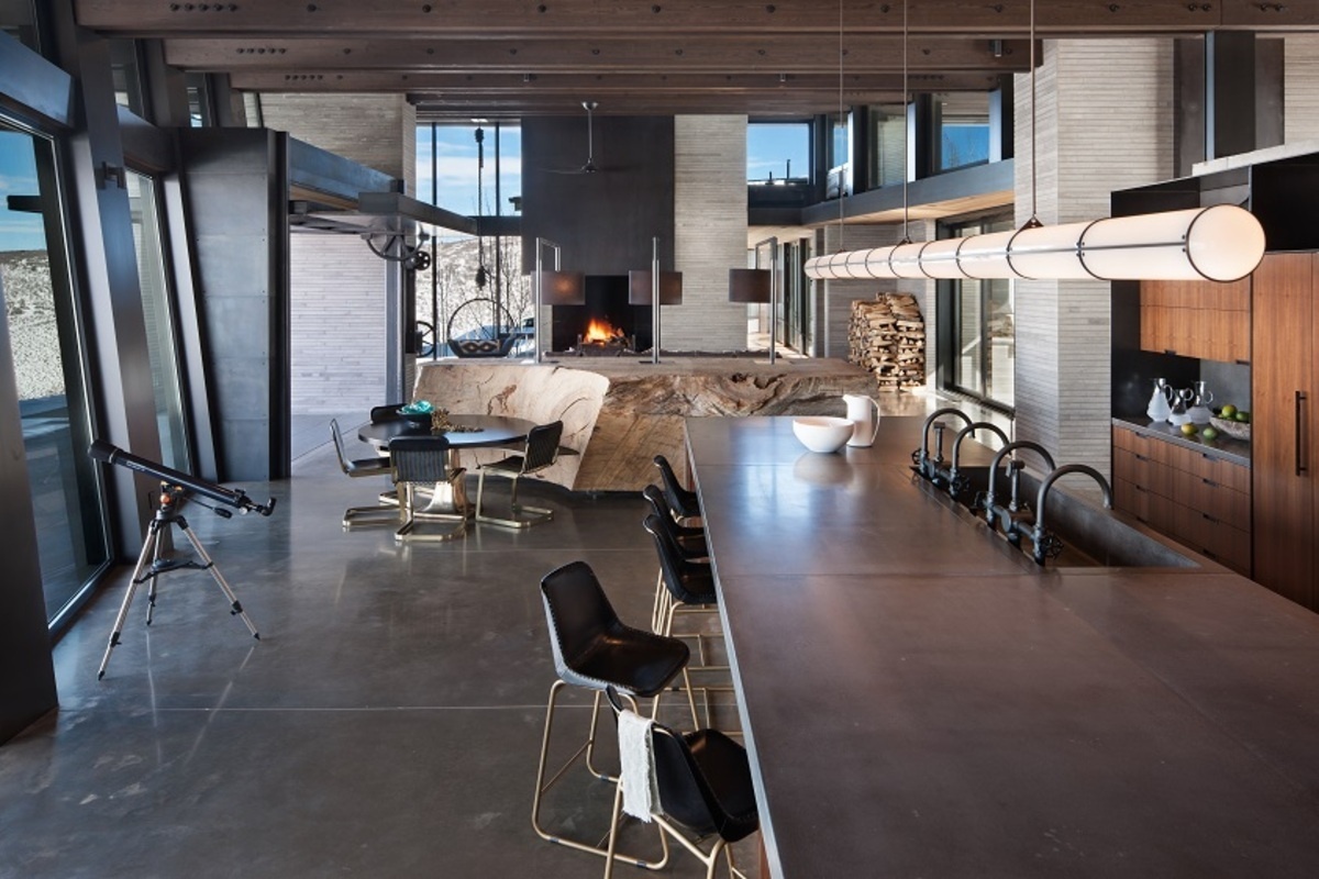 The open-plan kitchen, dining area, and living room at Quarry House are all oriented toward panoramic mountain views. Concrete floors, a sculptural fireplace hearth, built-in window seating, and a hanging chair create a relaxed yet sophisticated living space.