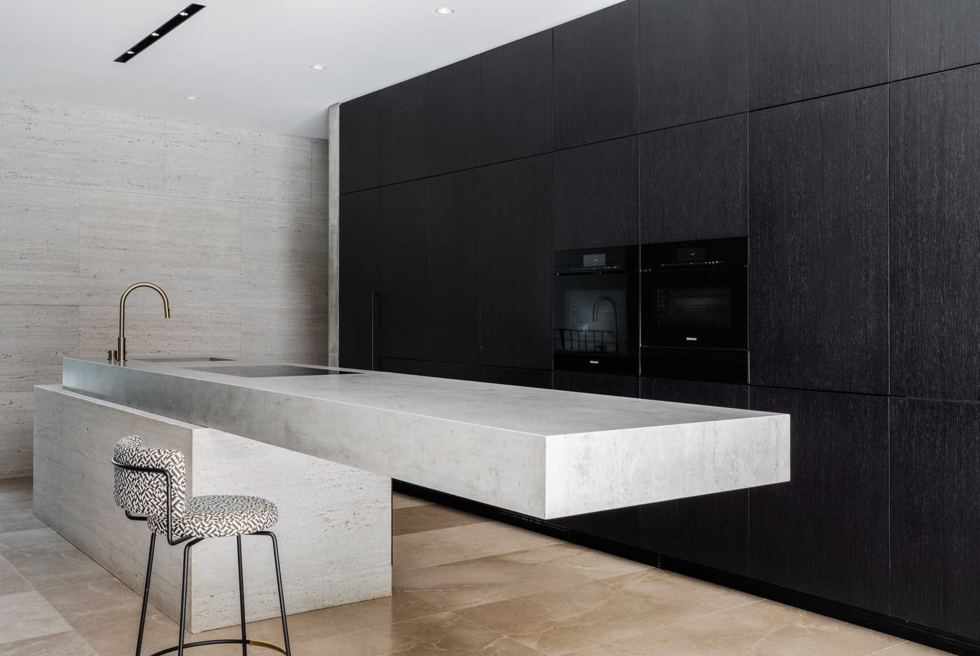 The minimalist kitchen in this Sydney home features a dramatic cantilevered island, black cabinetry, concrete walls, limestone floors, and travertine finishes that create a bold yet refined modern aesthetic.