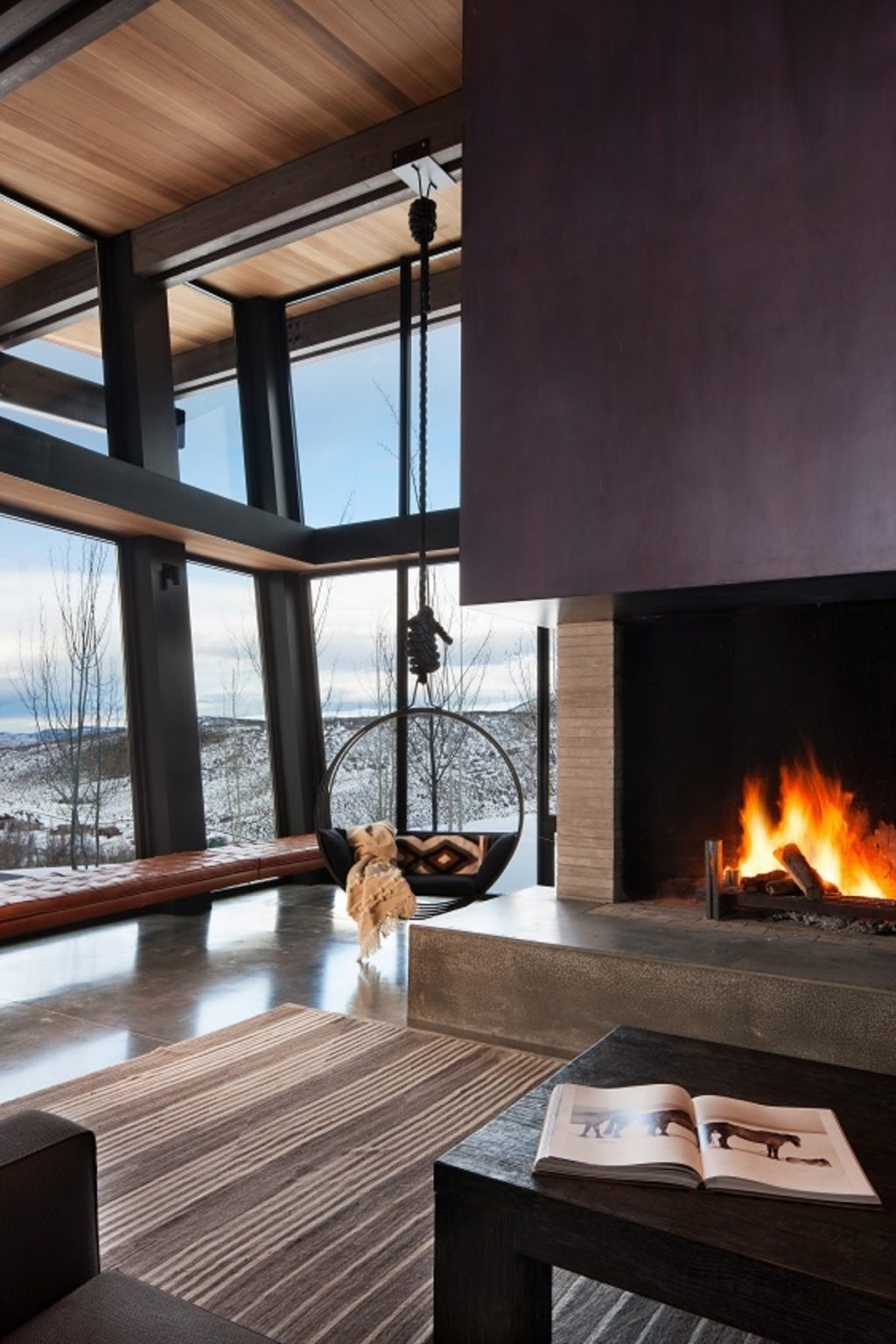 The open-plan kitchen, dining area, and living room at Quarry House are all oriented toward panoramic mountain views. Concrete floors, a sculptural fireplace hearth, built-in window seating, and a hanging chair create a relaxed yet sophisticated living space.