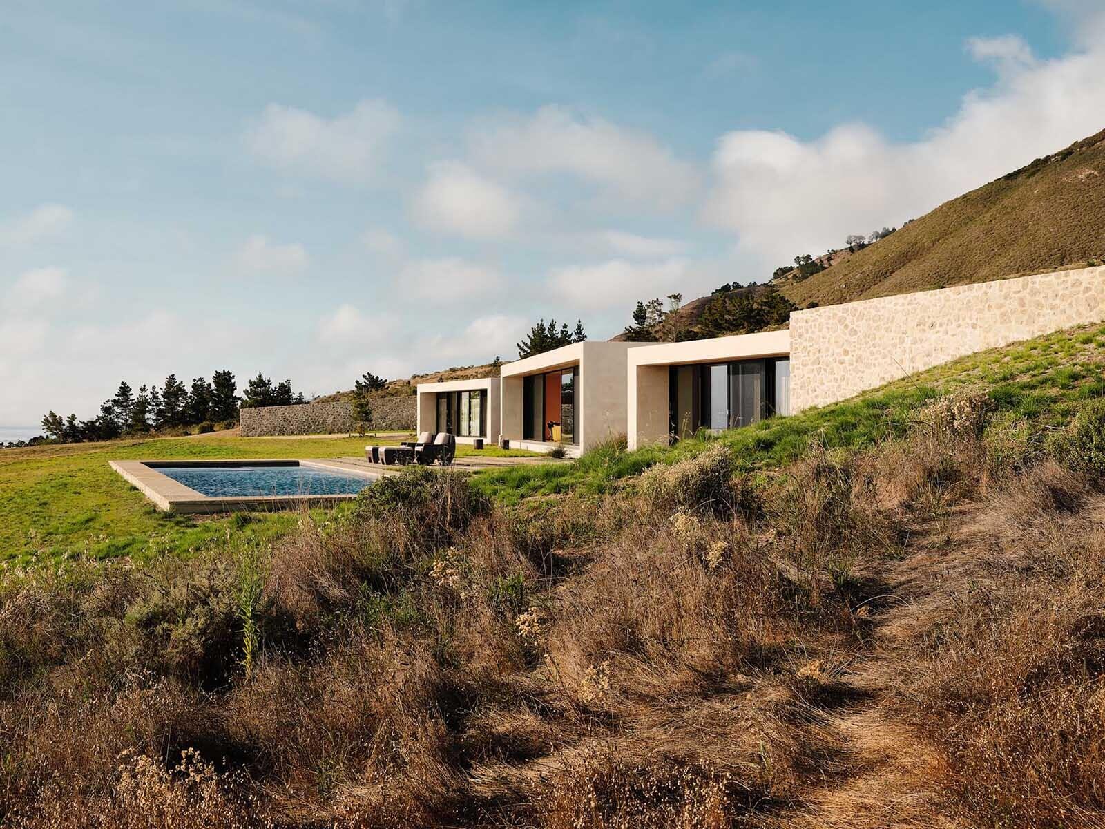 Stone walls, earth-toned concrete, and green roofs help this Big Sur home disappear into the hillside while floor-to-ceiling glass opens every space toward the Pacific Ocean.